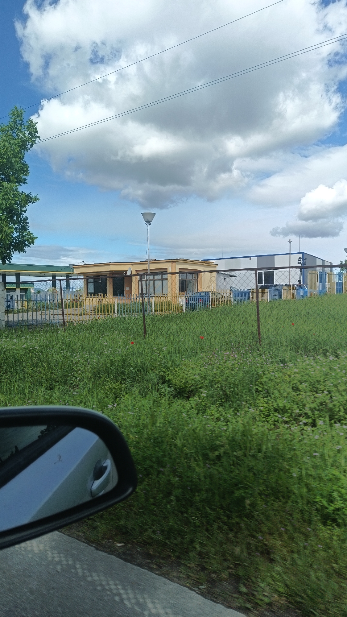 The image shows a view from a moving vehicle, specifically the driver's side mirror, with the roadside visible. The location appears to be in the city of Boliartsi, Bulgaria, based on geographical clues. In the midground, there are two buildings of distinct architectural styles, one light yellow and one grey and white, with fences surrounding them. A grassy field separates the road from the buildings, and some scattered red wildflowers are visible. Above, the sky is partly cloudy with a few overhead power lines. The vehicle is travelling on an asphalt road.