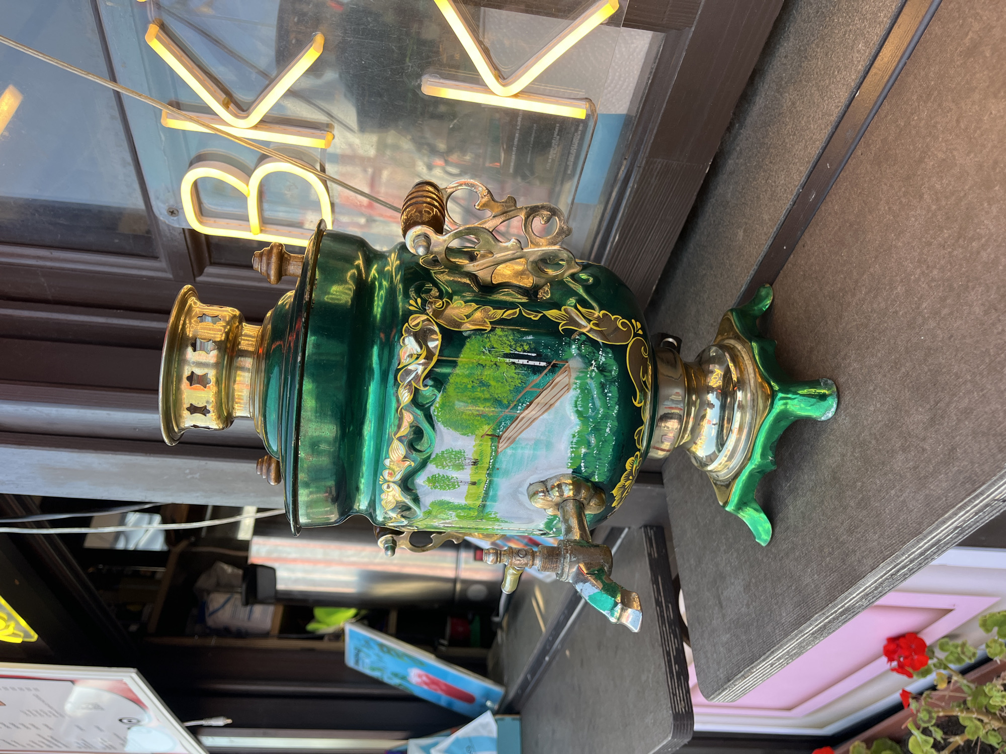 An ornate, emerald green samovar with gold trim and a painted landscape is displayed on a dark wooden shelf. The samovar is centrally placed and appears to be in focus. To the right of the samovar, a window reflects yellow neon letters that appear to spell "BK". Behind the samovar, a shelf in the background holds various items, including bags and what looks like a menu board with a picture of a red drink. The setting appears to be outdoors, possibly on a street or a cafe's exterior. The image is likely taken in Nizhny Novgorod, Russia, given the presence of a samovar, a traditional Russian item.