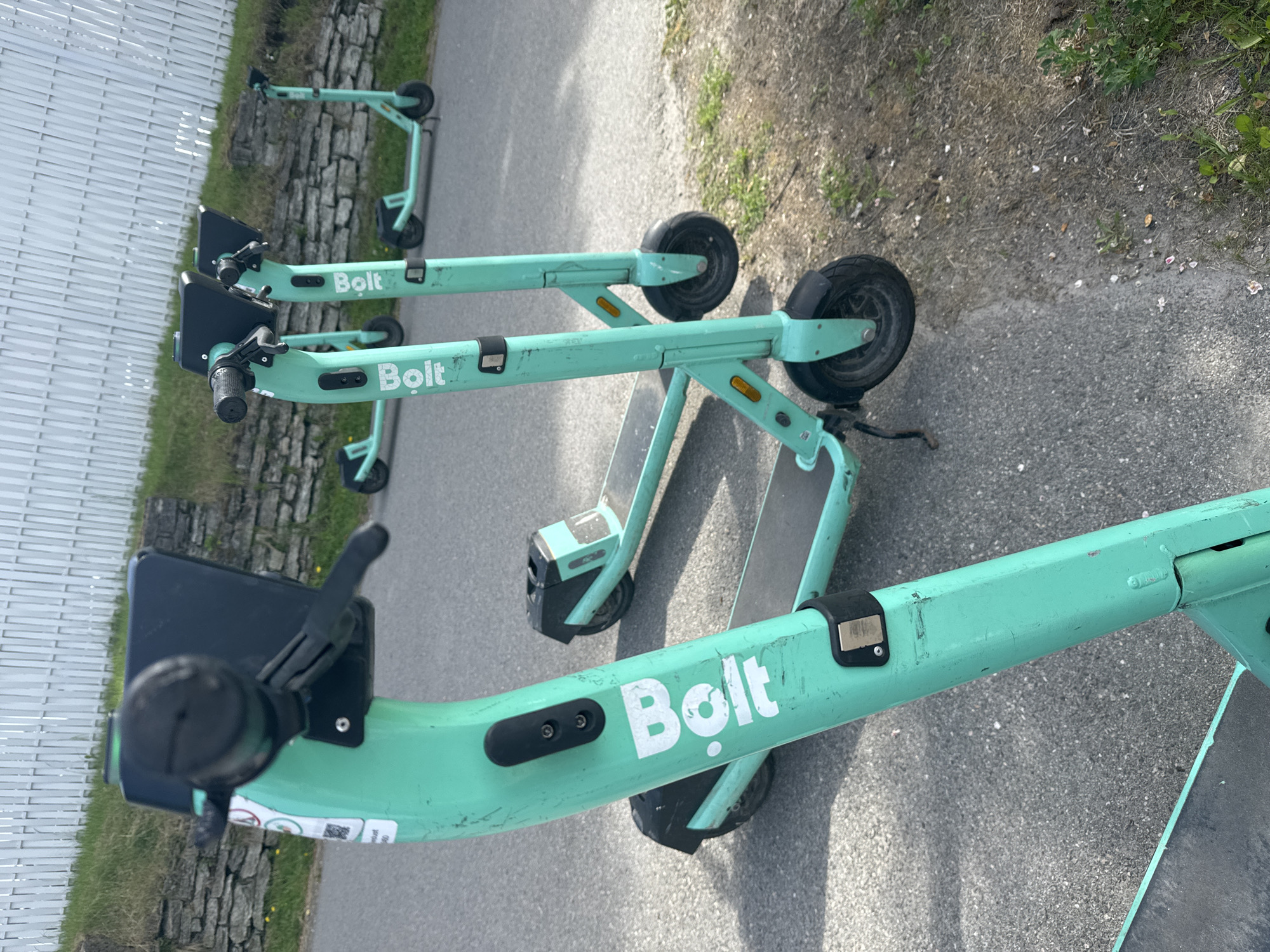 The image shows a row of green "Bolt" branded electric scooters parked on a paved path next to a grassy area with a stone wall and a white fence in the background. The scooters are lined up, with one in the foreground partially obscuring the others. They are all upright, suggesting they are parked and available for use. The environment appears to be an outdoor public space, possibly a park or a sidewalk, in Tallinn, Estonia. The lighting indicates it is daytime. There are no people visible in the image. The condition of the scooters appears to be used, with some scuff marks and wear visible on their surfaces.