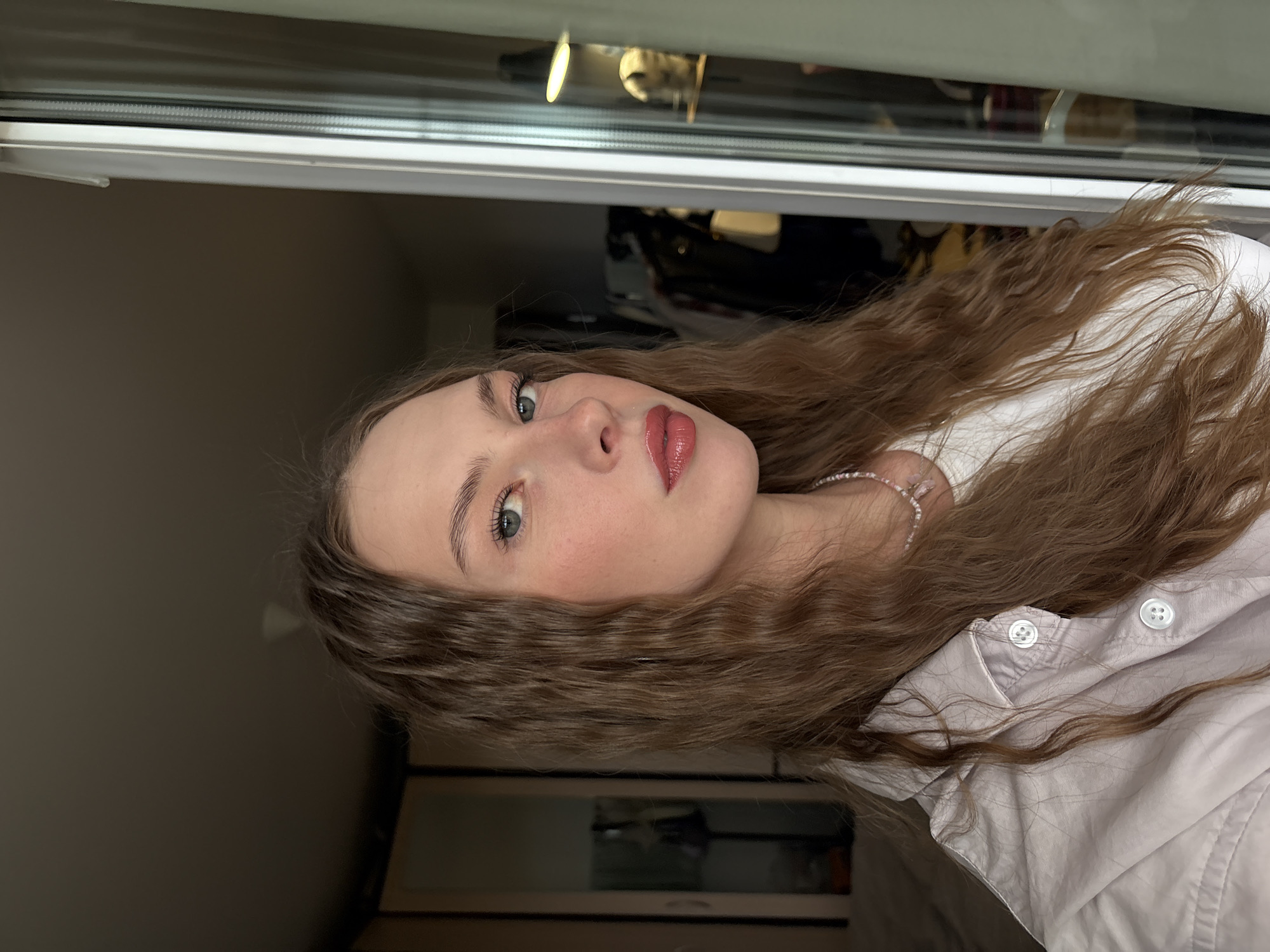 A young Caucasian woman with long, wavy brown hair is captured in a close-up, selfie-style portrait. She is wearing a light-colored collared shirt over a white t-shirt and a beaded necklace. Her makeup includes blush and reddish-pink lipstick. The background is a room with dark walls, featuring a wardrobe with mirrored doors on the left and a window with sheer curtains on the right. The lighting suggests an indoor setting, possibly a bedroom. The location is the city of Perm, Russia.