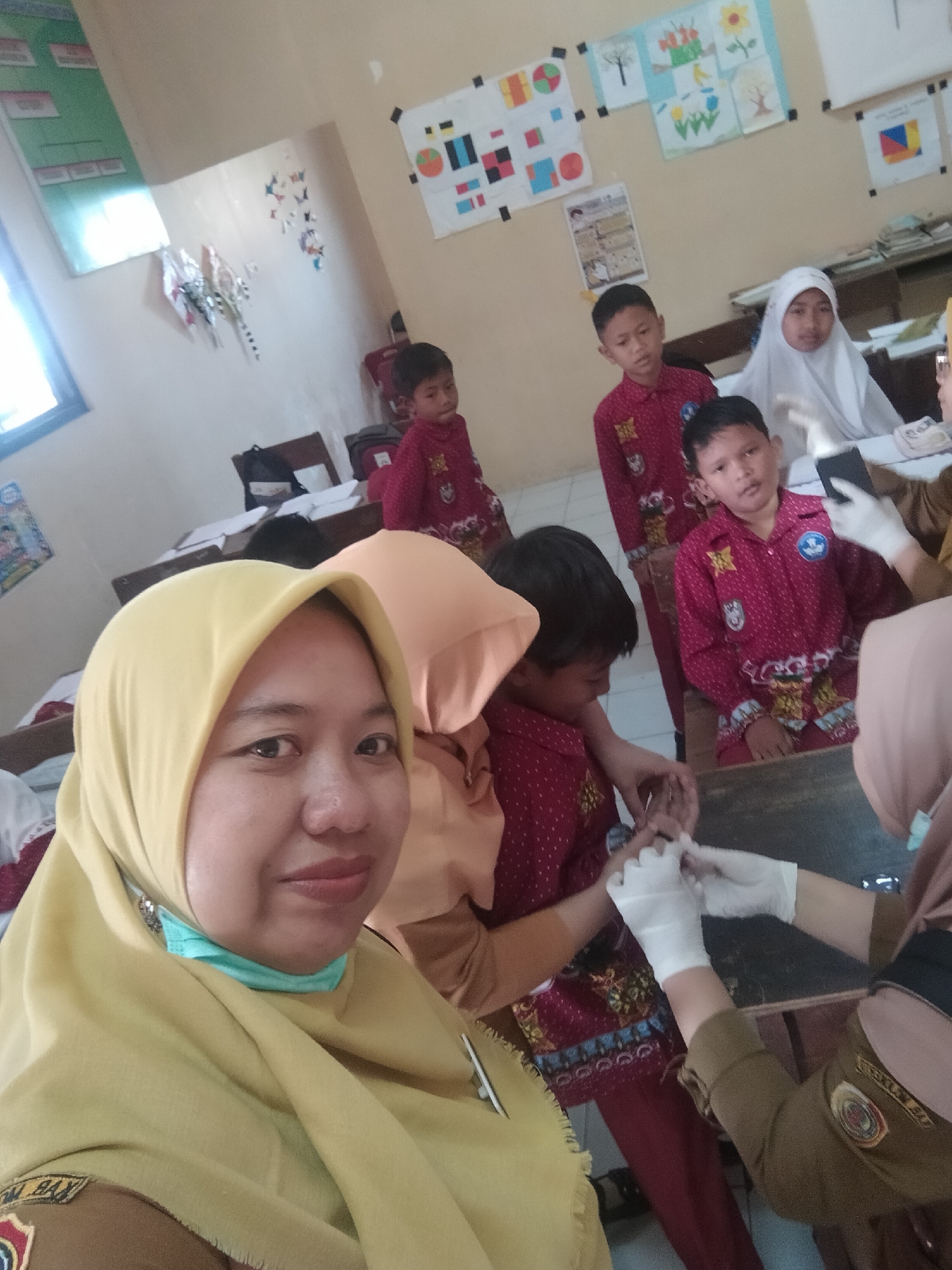 The image depicts a classroom setting in Tretes, Indonesia, possibly at a school, as evidenced by the educational posters on the wall and the presence of children in uniforms. A woman in a yellow hijab is in the foreground, smiling at the camera. Behind her, several children in red uniforms with patterned accents are present. One of the children is receiving medical attention, with an individual wearing white gloves tending to their hand. Another child, wearing a white hijab, is seated to the right. The room appears to be a typical classroom with desks and chairs. The overall atmosphere seems to be one of a school activity or event.