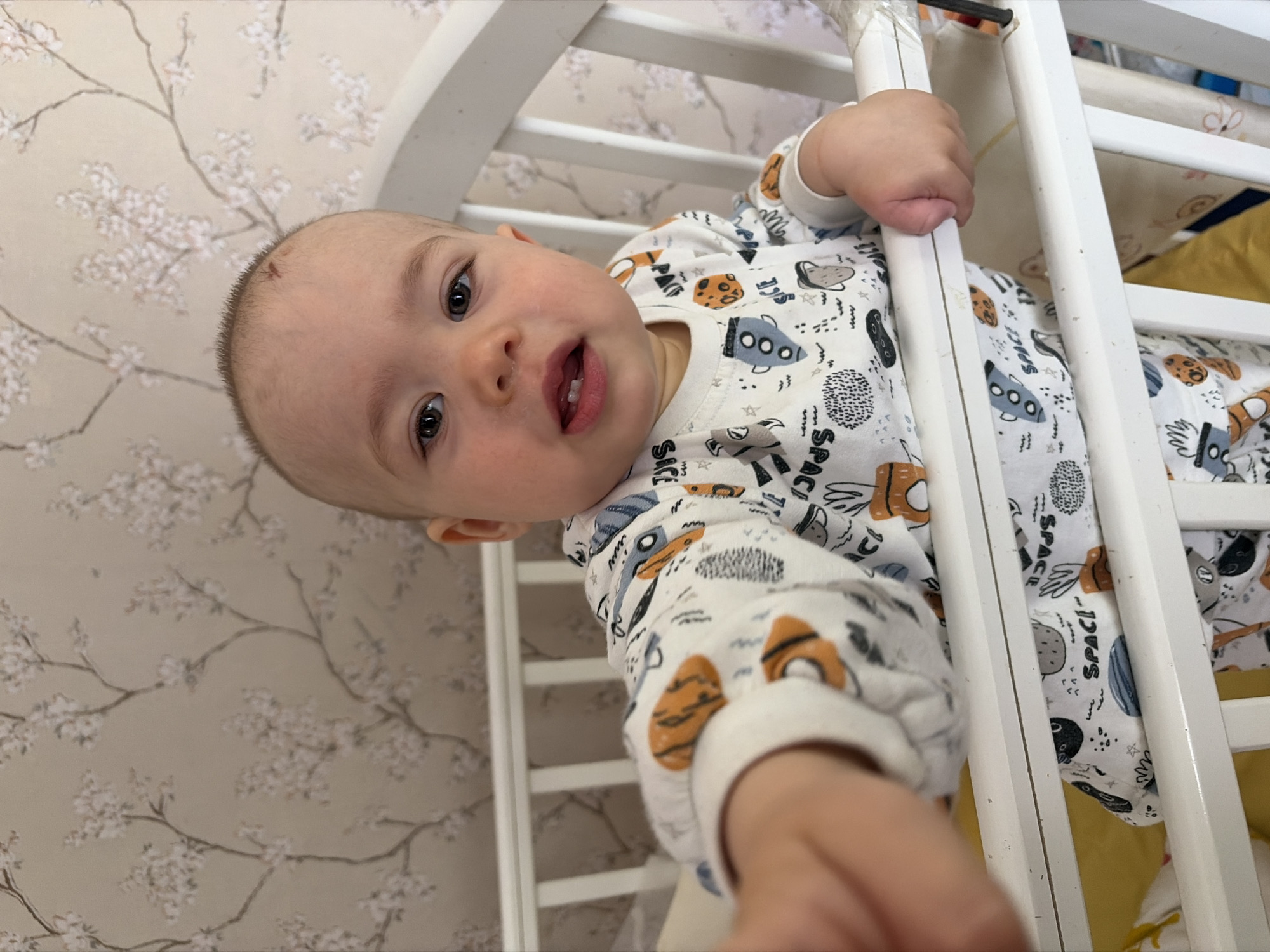 A baby is lying in a white crib in a room in Chaikovsky, Russia. The baby is wearing a white pajama top printed with rockets, planets, and the word "SPACE." The baby is holding onto the side of the crib with their hands. The crib has a rounded headboard. Behind the crib is a wall covered with a wallpaper pattern of light gray flowers. The crib mattress has a yellow and white pattern.