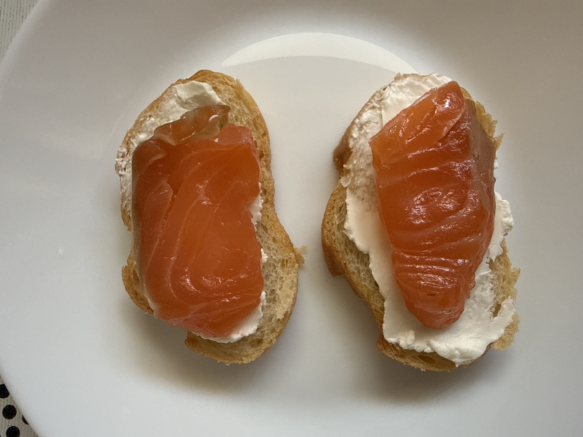 The image shows two small pieces of bread, each topped with a layer of white cream cheese and a piece of orange-colored salmon. They are placed on a white plate. The scene is indoors, and the location is unspecified.