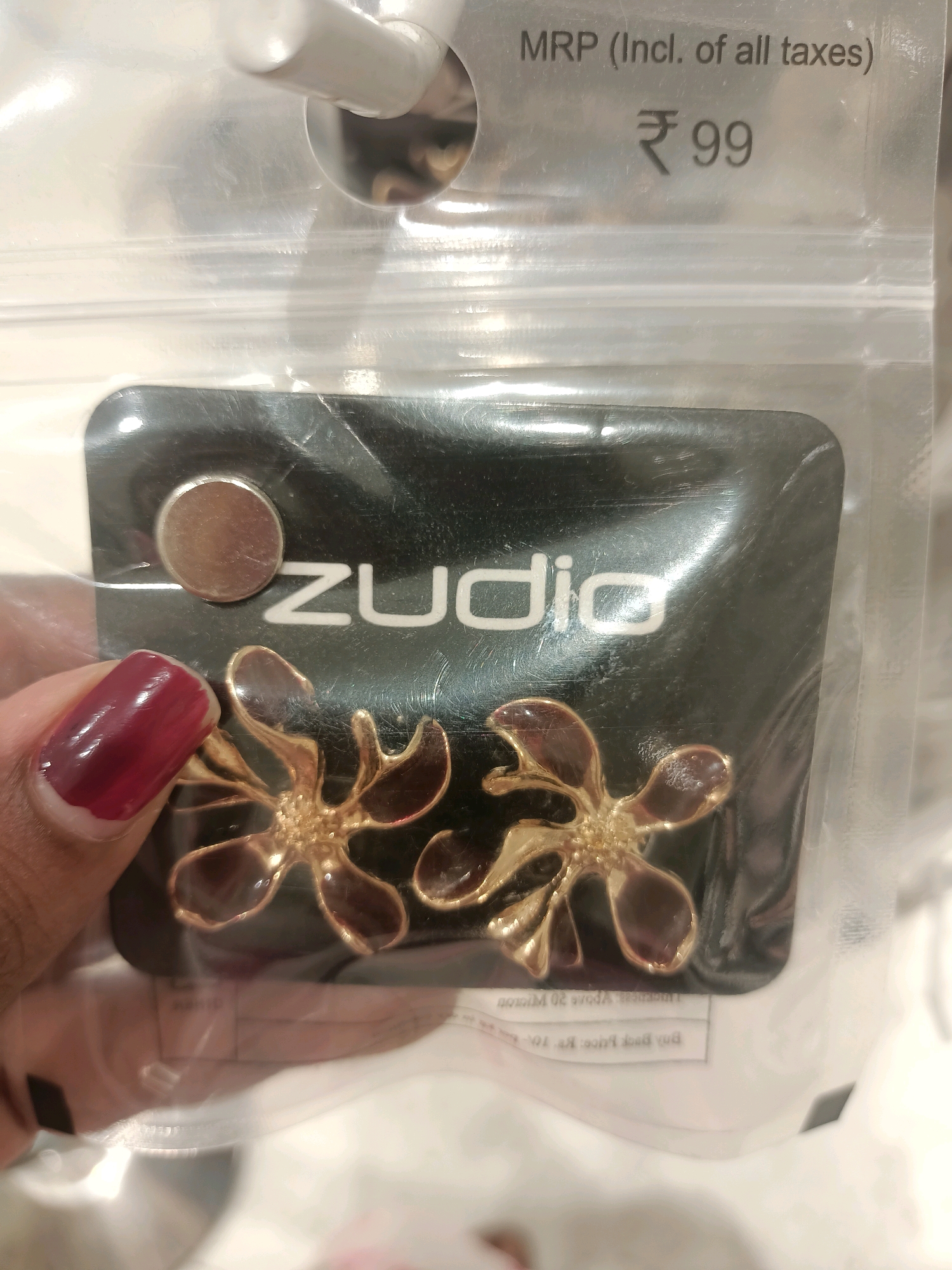 A close-up shot shows a pair of gold-toned flower-shaped earrings packaged in clear plastic. The earrings are attached to a black card featuring the white text "zudio". Above the earrings, the price is displayed as "MRP (Incl. of all taxes) ₹ 99". A person's hand with dark red nail polish is visible in the lower left corner, holding the packaging. The background is blurred, but appears to be indoors, possibly a retail environment. There is no information in the image that can be used to determine a specific location within Barrackpore, India.