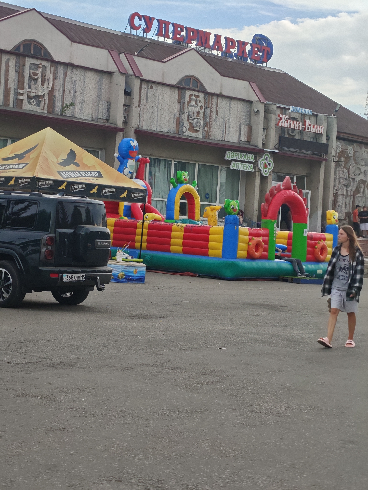In Petropavl, Kazakhstan, a black Haval SUV is parked on an asphalt surface in front of a building. The building has a sign with Cyrillic lettering that reads "СУПЕРМАРКЕТ" (Supermarket) in red on its facade. To the left of the SUV, there is a yellow tent with a logo featuring two eagles. Adjacent to the tent, a colorful inflatable playground with cartoon characters is set up. To the right of the playground, a young woman wearing a plaid shirt, gray shorts, and pink slides walks away from the viewer. The sky above is partly cloudy.