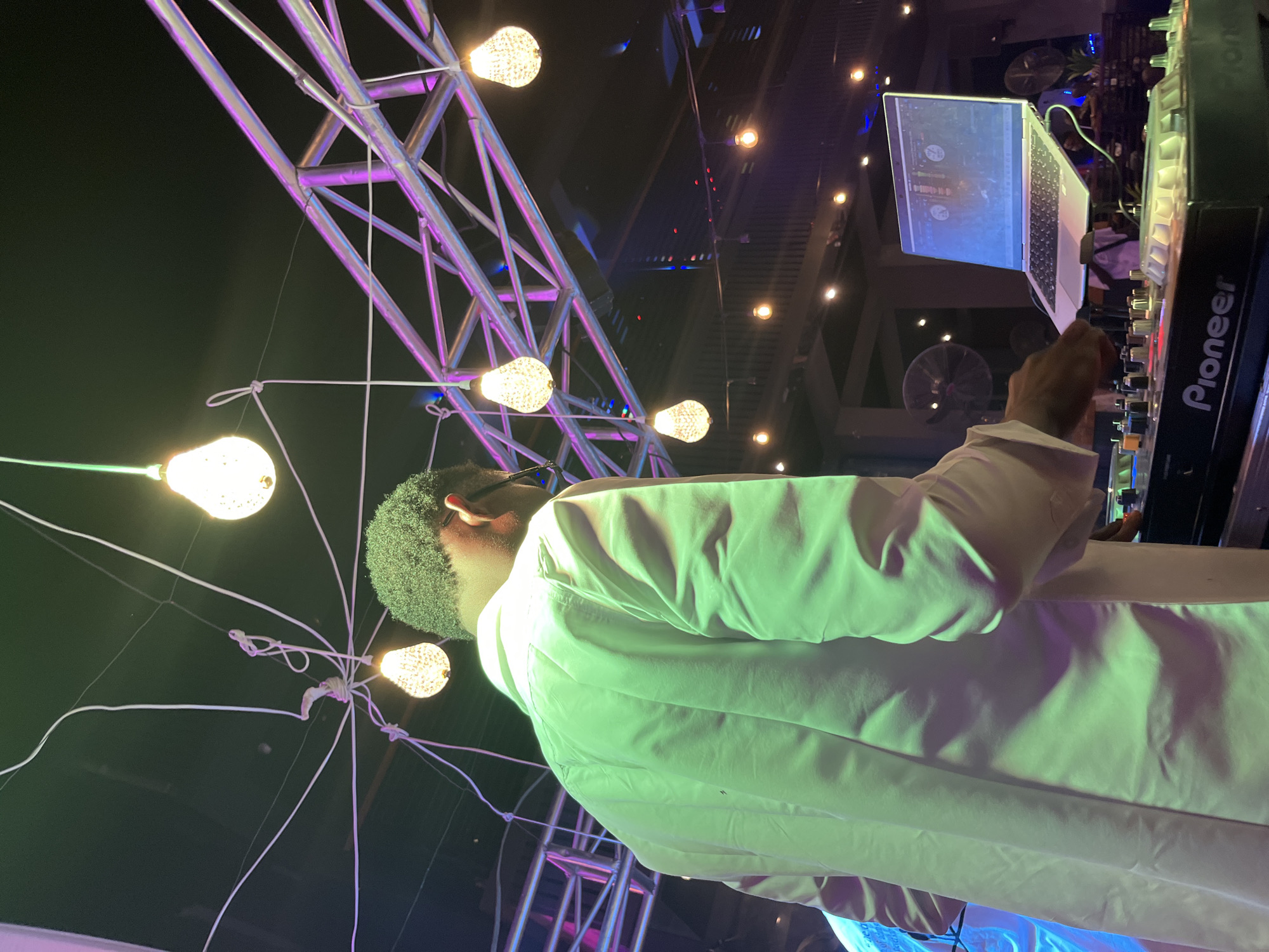 The image depicts a person, appearing to be a DJ, operating a DJ setup at what seems to be an outdoor event at night. The DJ is wearing a light-colored, possibly white, long-sleeved shirt and is facing away from the camera, towards the DJ equipment. The DJ setup includes a laptop placed on top of a Pioneer DJ mixer. The laptop screen is illuminated and displays what appears to be DJ software.
The scene is decorated with numerous string lights, some of which resemble Edison bulbs and are illuminated, casting a warm glow. These lights are suspended by thin wires, creating a web-like structure. In the background, a large, metallic truss structure is visible, illuminated with purple and pink lights, suggesting a stage or performance area. The overall lighting in the scene is dynamic, with the purple and pink lights from the truss contrasting with the warm light from the string bulbs and the blueish light from the laptop screen.
The time of day is clearly night, as indicated by the dark sky and the prominent artificial lighting. No specific weather conditions are discernible. The location appears to be an open-air venue, possibly a garden, plaza, or outdoor event space, given the truss structure and the string lights. The atmosphere seems festive and energetic, typical of a music event.
Visible text includes the brand name "Pioneer" on the DJ mixer. The DJ's hair is curly and dark. The image is captured from a slightly low angle, looking up towards the DJ and the overhead lighting.