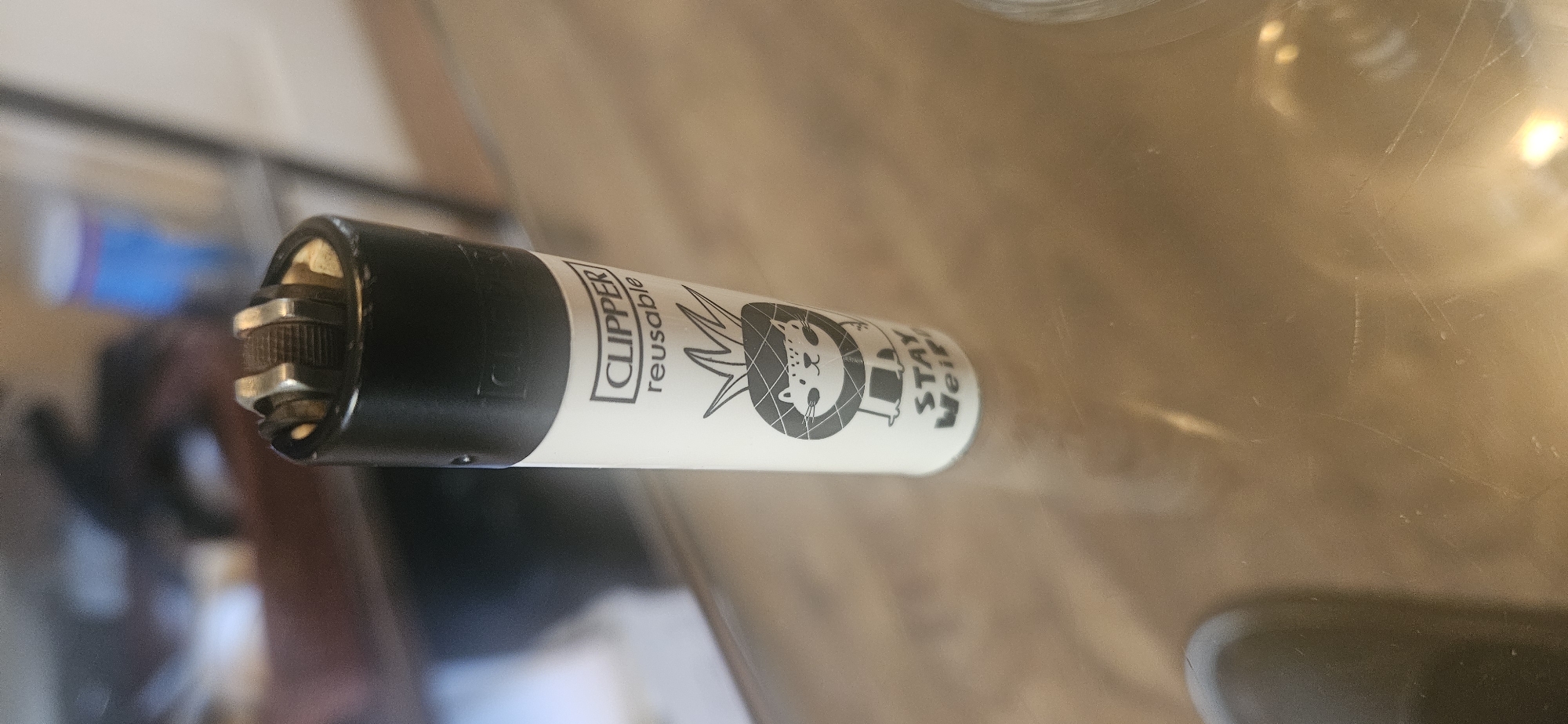 A Clipper brand lighter, with the words "CLIPPER reusable" and a black-and-white drawing of a lion and the words "STAY Weird" on the side, is lying horizontally on a reflective surface. The top of the lighter is black, and the body is white. The surrounding environment is blurry, but appears to be indoors. The location in the city of Norwich, United Kingdom cannot be identified from the image.