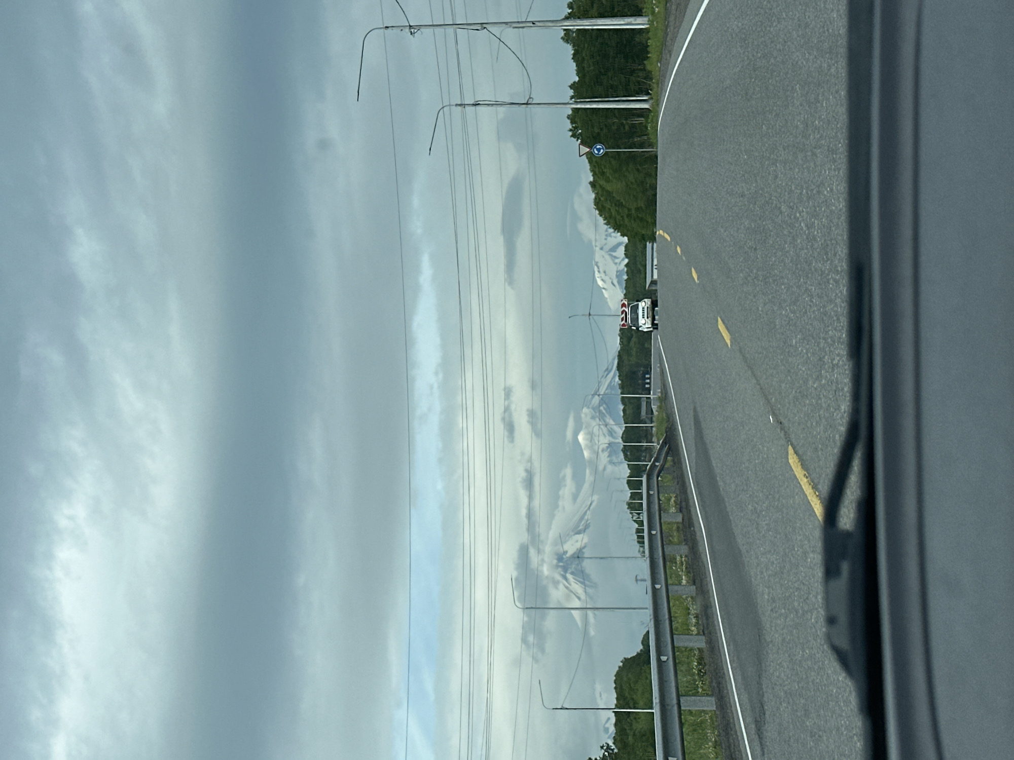 The image shows a view from inside a vehicle driving on a road with snow-capped mountains in the distance. The sky is overcast with thick, grey clouds. Power lines run horizontally across the upper portion of the image. A metal guardrail is visible along the left side of the road. In the middle distance, a white vehicle with red and white signage on its roof is driving in the same direction as the camera. Trees line the right side of the road, and a traffic sign with a blue circle and white arrows is visible. The road is paved and has yellow and white lane markings. The foreground shows the dashboard and windshield of the vehicle from which the photo was taken. The location is identified as the city of Krutoberegovy, Russia.