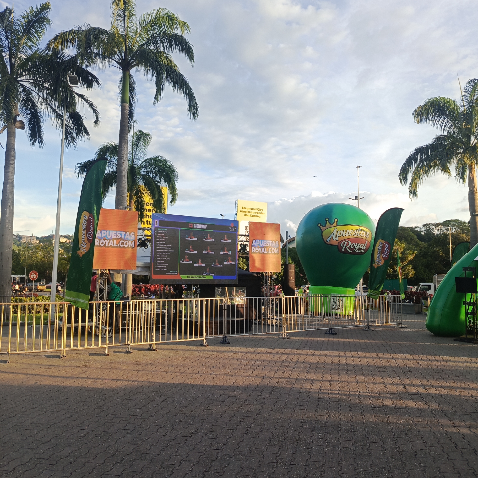 The image depicts an outdoor scene in Caracas, Venezuela, likely during a sporting event or promotion. In the foreground, a metal barricade separates the viewer from a paved area where several promotional elements are set up. Dominating the scene are large green inflatable balloons with the "Apuestas Royal" logo, one of which is a giant sphere and another a curved structure. To the left, a large LED screen displays what appears to be a FIFA World Cup roster with player names and images, indicating a sports-betting promotion. Several flags with the "Apuestas Royal" logo are also visible, as is an orange banner with "APUESTAS ROYAL.COM" printed on it. Palm trees frame the top of the image, and in the background, buildings on a hill suggest the urban landscape of Caracas. A few people are visible near the promotional setup, some appearing to be staff. The lighting suggests it is late afternoon or early evening, with long shadows cast across the paved ground.