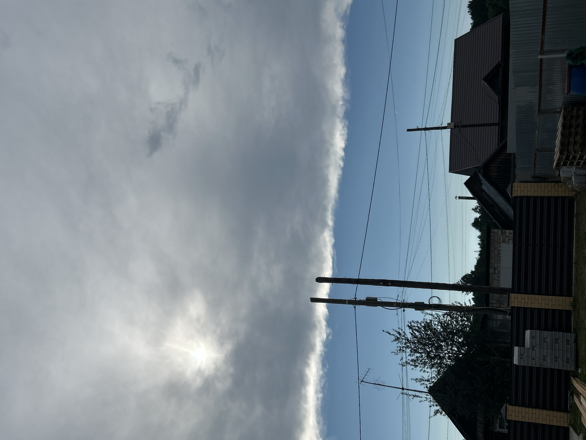 This image depicts a residential area in Chaikovsky, Russia. The sky is partly cloudy with a prominent, long band of gray clouds stretching horizontally across the mid-ground. Above this band, the sky is brighter, with the sun partially obscured by thinner clouds, creating a diffused light. Below the gray cloud band, the sky is a clear blue.
In the foreground, there is a dark brown corrugated metal fence with brick pillars. To the left of the fence, a stack of gray concrete blocks is visible. Beyond the fence, there are several houses with dark-colored roofs, typical of rural or suburban Russian architecture. One house on the left has a dark green, dense tree in front of it. Two tall wooden utility poles stand near the center of the image, with wires strung between them and other unseen points. A TV antenna is visible on a pole to the left. In the background, a line of dark green trees forms a horizon. The overall impression is that of a quiet, residential neighborhood, possibly on the outskirts of the city, with overcast conditions beginning to clear.