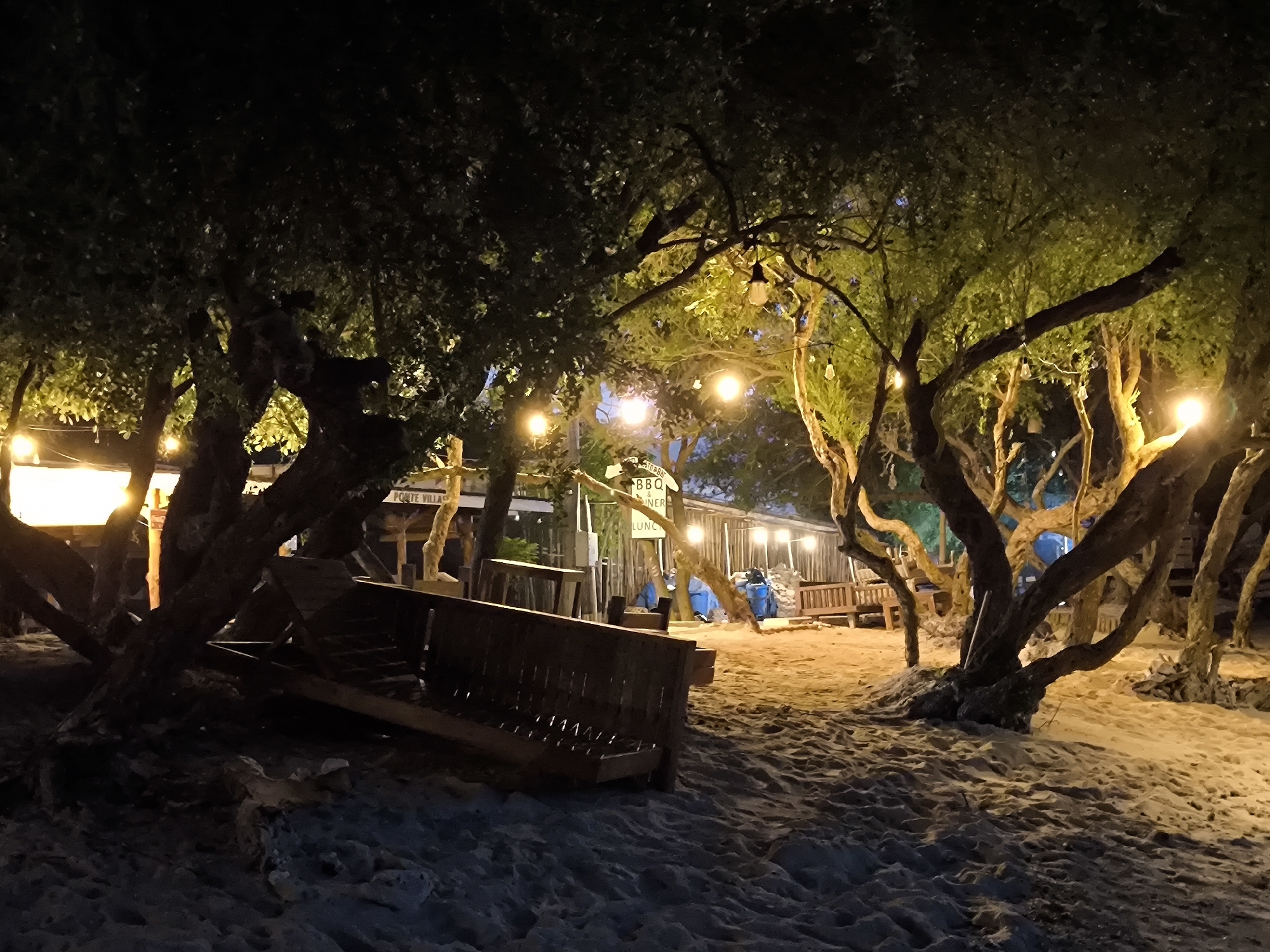 The scene is a beach at night, with sand covering the ground and a few trees scattered around. String lights are hung between the trees, providing illumination. A wooden bench sits on the sand. In the background, a sign reading "BBQ DINER LUNCH" is visible. The location is Unknown, Indonesia. No people are visible.