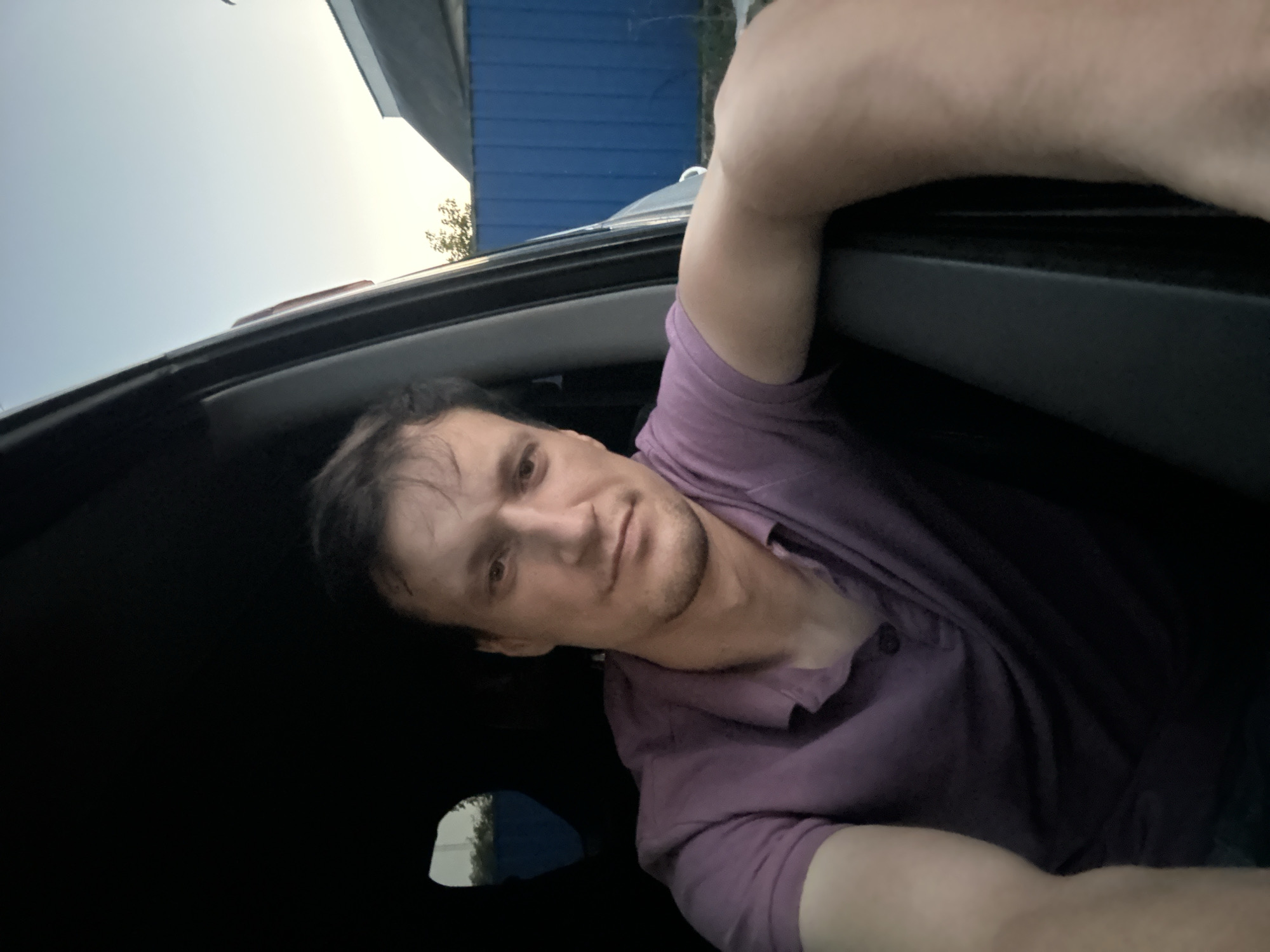 In Shatava, Ukraine, a man is captured in a selfie taken from inside a vehicle. He is seated in the driver's seat, with his head and upper torso visible. The man has dark, disheveled hair and is looking directly at the camera with a neutral expression. He is wearing a purple polo shirt. His right arm is resting on the car door frame, and his left arm is also visible, partially obscuring the frame. The interior of the car is dark, with a small window on the left revealing a glimpse of greenery. Outside the car, a blue wooden fence and the roof of a building are visible under a pale sky. The overall lighting suggests it is either early morning or late evening.