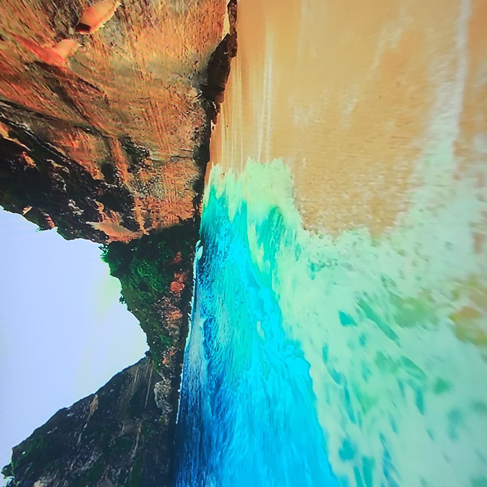 The image shows a coastal scene with a beach, ocean, and cliffs. The cliffs are on the left side of the image, displaying a combination of rock formations and vegetation. The beach is on the right, meeting the turquoise-colored water. The ocean water has a foamy appearance, suggesting wave action. The sky is visible in the top left corner, appearing light blue. The image is rotated counterclockwise. The image is not of Yerevan, Armenia.