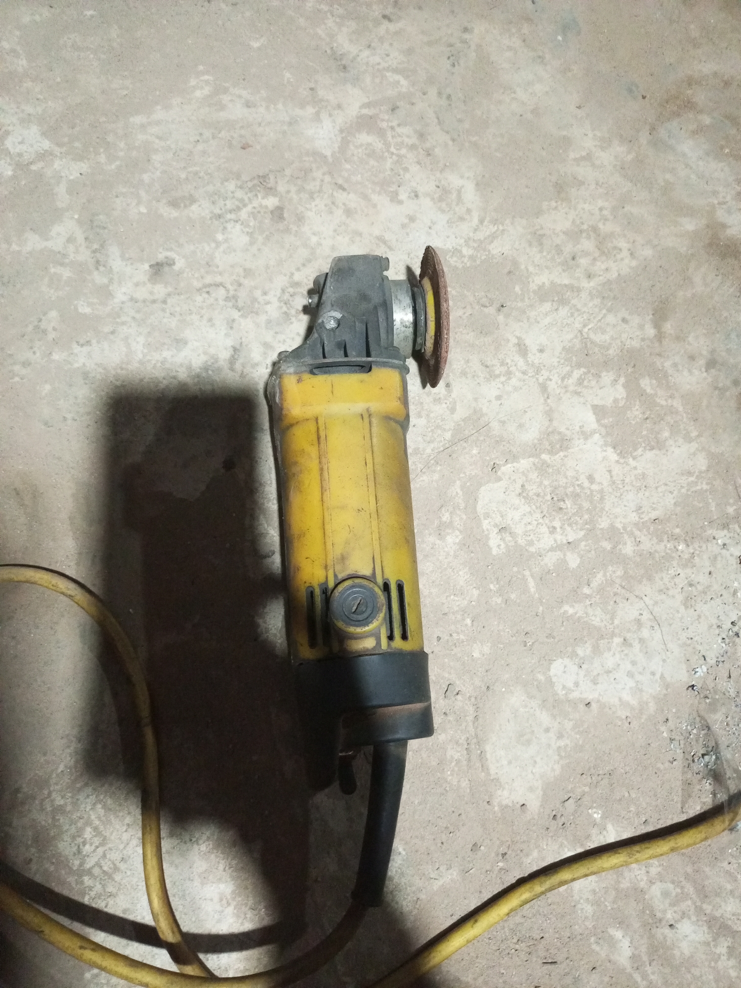 An angle grinder with a yellow body and a cutting disc is lying on a concrete floor in the city of Keling Panggau, Indonesia. The grinder is connected to a yellow power cord that snakes across the floor. A shadow of the grinder is cast on the concrete to its left. The concrete floor appears rough and uneven with some debris scattered around. The lighting suggests an indoor environment with overhead light sources.