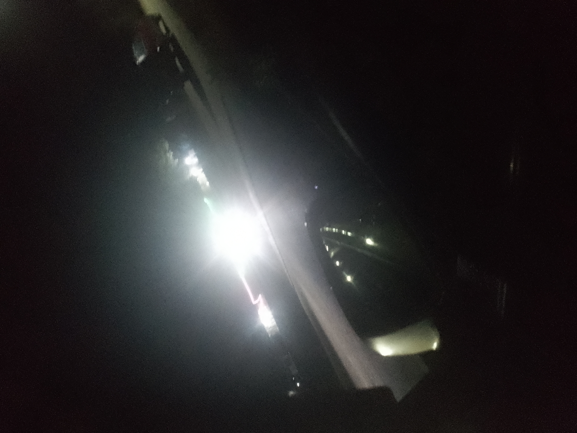 The image depicts a dimly lit outdoor scene, likely at night, with strong artificial lighting obscuring much of the detail. In the foreground, a white, bright light source dominates the left side of the frame, causing lens flare and overexposure. To the right of this light, a blurry, reflective surface, possibly a car windshield or window, is visible, angled upwards. Beyond this surface, faint, scattered lights suggest the presence of buildings or streetlights in the distance. The overall impression is one of a poorly lit urban environment at night. The specific location in Jalingo, Nigeria cannot be determined from the visual information provided.