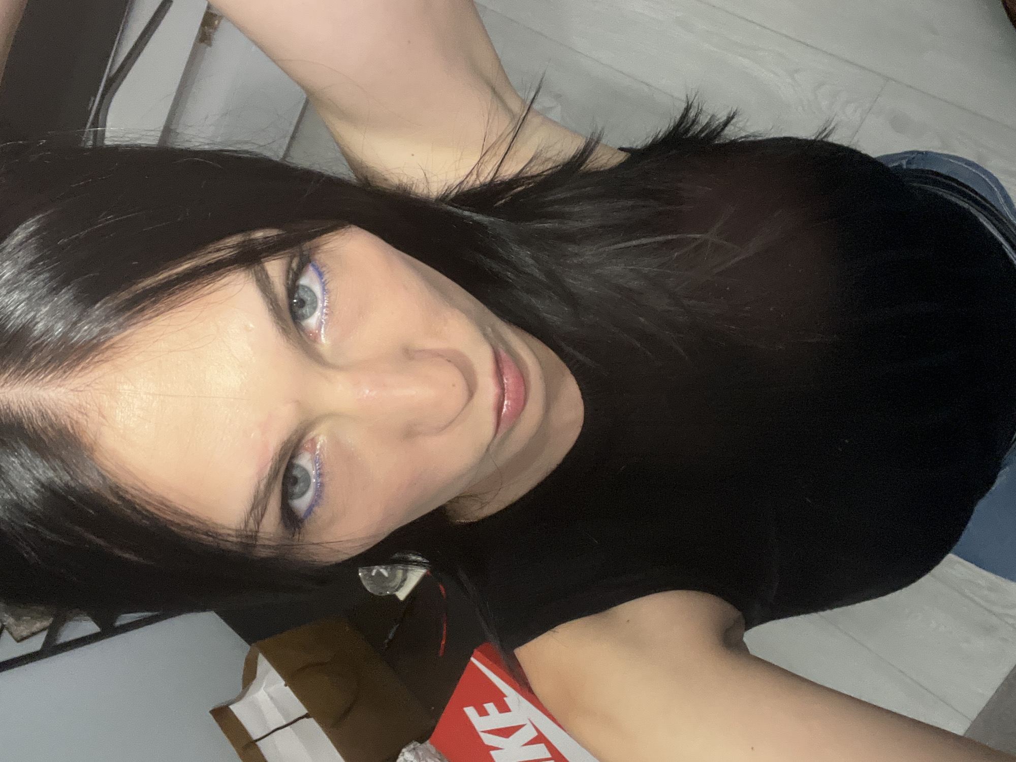 An overhead, slightly angled shot captures a young adult female looking directly into the camera. She has long, dark hair, blue eyes accentuated with purple eyeliner on the lower lash line, and glossy pink lips. Her expression is neutral to slightly smiling. She is wearing a black t-shirt, and a section of blue denim fabric, likely from jeans, is visible on her lower right. Her right arm is visible, bent at the elbow, positioned above her head.
The setting appears to be indoors, with a light-colored, possibly wooden, floor forming the background. In the lower left of the frame, a red and white box or packaging is visible, displaying part of the Nike swoosh logo and the letters "KE." Other indistinct dark shapes and furniture elements can be seen in the background. The primary activity is the subject posing for the camera. There are no clear indicators of the time of day or specific weather.