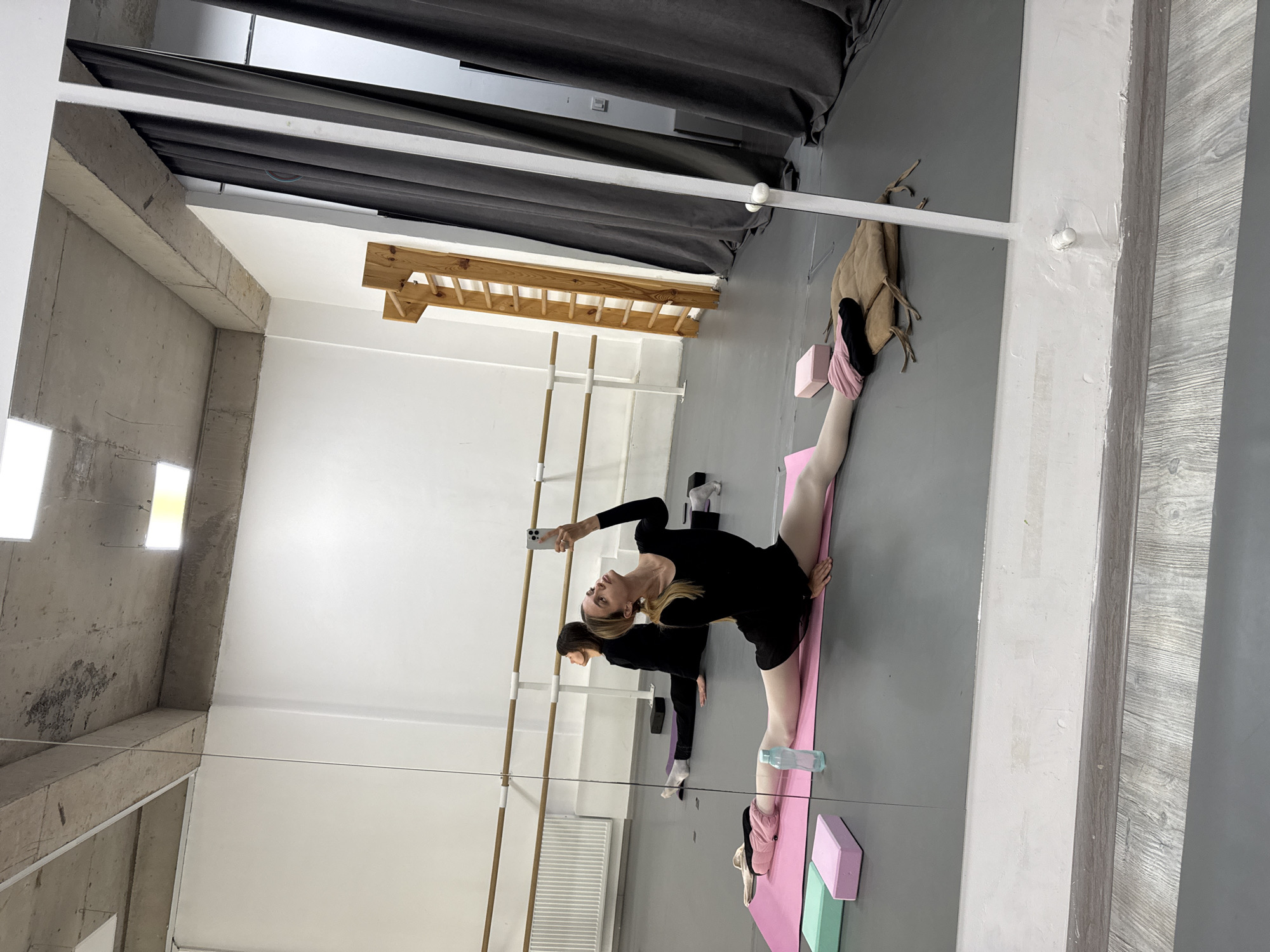 A mirrored room contains two ballet barres, light gray flooring, and concrete walls. A ladder-like structure hangs from the ceiling. Gray curtains are positioned overhead.
In the foreground, a ballet dancer in a black leotard is doing the splits on a pink mat, using pink and mint foam blocks. She holds a cell phone, taking a picture of herself in the mirror. Another dancer, also in black, is stretching near the ballet barre.
A bag hangs from the mirror on the right.
The location of this image is Tbilisi, Georgia.
