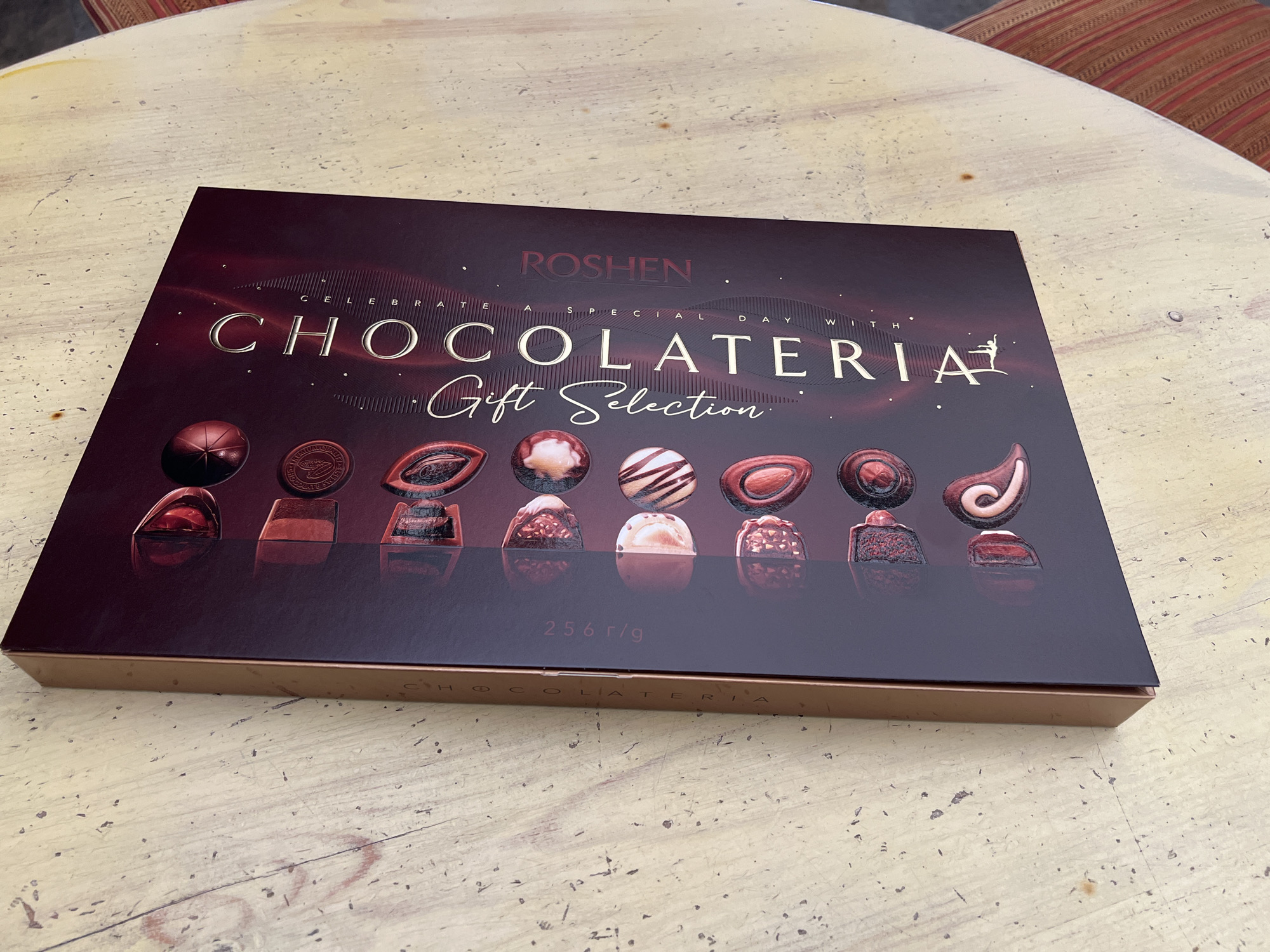 A close-up, top-down shot shows a dark maroon gift box of chocolates, labeled "CHOCOLATERIA GIFT SELECTION" in gold lettering. The box is positioned at an angle on a light-colored wooden surface, possibly a table, with visible grain and scattered dark specks. The chocolates themselves are arranged in a grid on the box lid and are depicted with various shapes and textures, rendered in shades of red and brown. The background is blurred, hinting at a textured, possibly striped fabric to the right. The scene appears to be indoors or in a sheltered outdoor area. There are no people or discernible actions occurring. The overall impression is one of a product shot or still life arrangement. The provided information about Nesebar, Bulgaria, is not visually verifiable from the image.