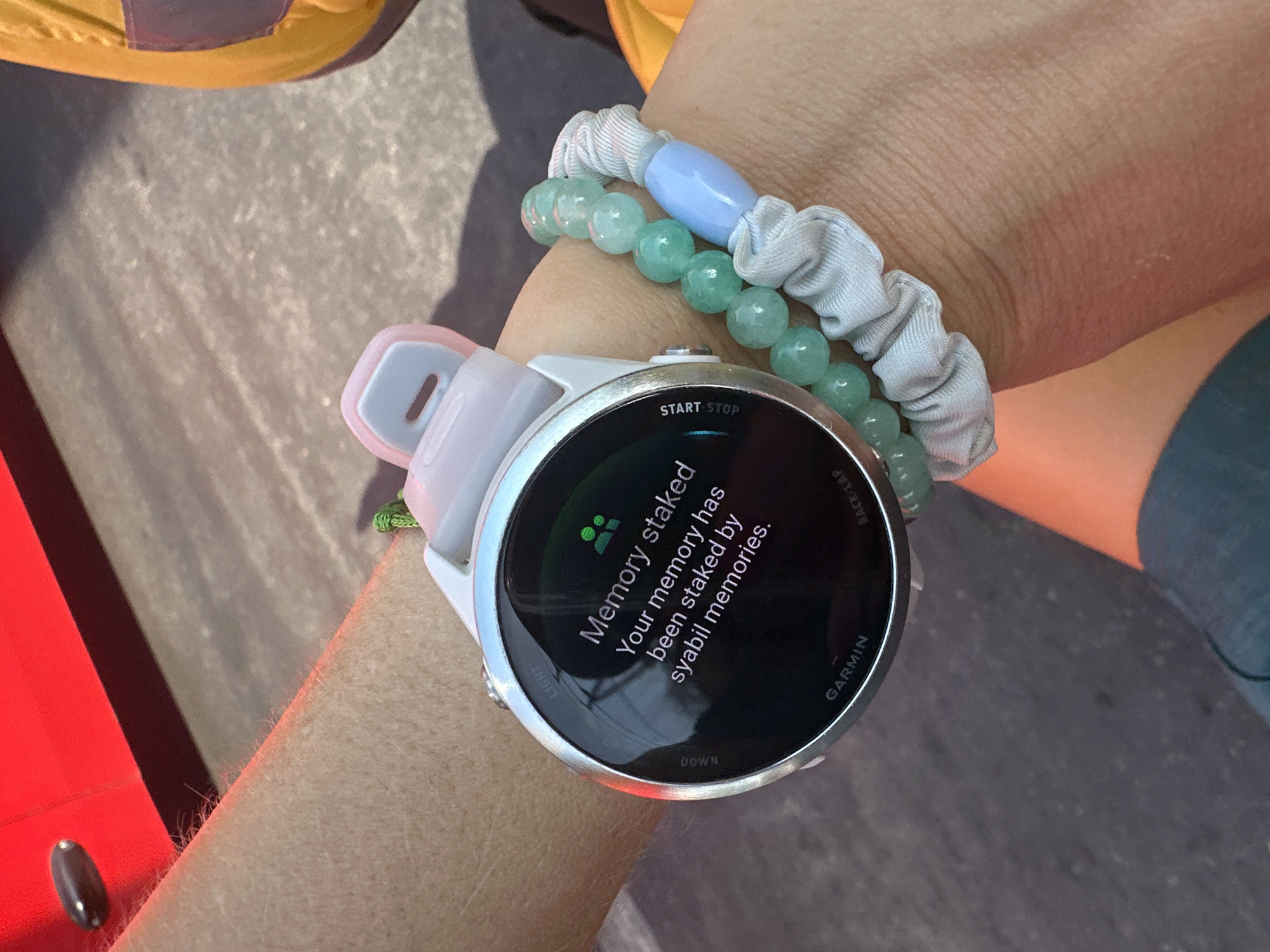 The image shows a close-up of a person's wrist wearing a Garmin smartwatch and two beaded bracelets. The smartwatch screen displays the text "Memory staked Your memory has been staked by syabil memories." The background is blurred and shows a grey, textured surface which could be pavement, and a bright red object with a dark shadow. There are also hints of yellow and purple in the background, possibly from clothing or another object. The scene appears to be outdoors. There is no visible indication of the location being Bac Nha Trang Ward, Vietnam.