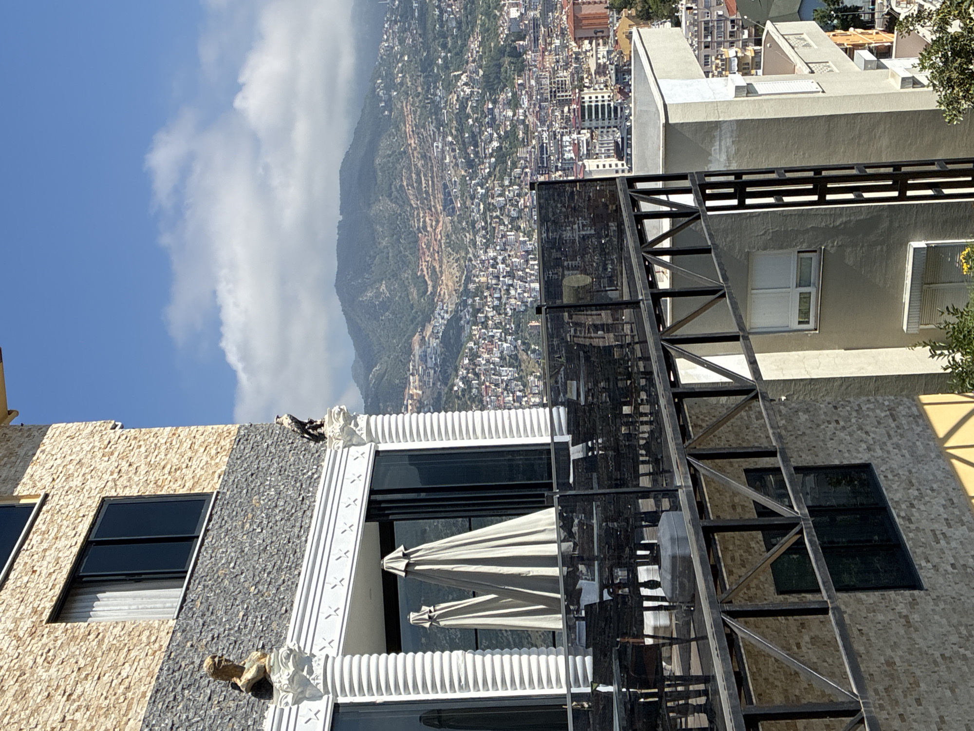 This media file presents a panoramic view from a high vantage point, looking down over a densely built city sprawling across a steep hillside. In the distance, lush green mountains rise under a bright blue sky dotted with soft white cumulus clouds.
In the foreground, several modern buildings are visible. The most prominent structure, located towards the bottom right, features a distinctive facade. It is constructed with two types of textured stone: a light beige on one section and a dark grey or black on another, separated by decorative white architectural elements. These include fluted columns, pilasters adorned with star patterns, and ornate carved corbels or gargoyles. This building features large, dark-paned windows. Extending from this structure is a contemporary balcony or terrace, equipped with sturdy dark metal railings and at least two closed patio umbrellas, suggesting an outdoor seating area. Another building with a similar stone facade is partially visible on the bottom left.
The midground and background are dominated by the extensive urban development, with countless multi-story buildings packed closely together along the contours of the hill. Beyond the city, the mountainous landscape stretches into the distance, with some peaks partially obscured by clouds.
The scene is captured during bright daylight hours, indicated by the clear visibility and distinct shadows cast by the buildings. There are no people visible, nor are there any discernible activities taking place. No text is visible within the image. The setting is identified as Alanya, Turkey.