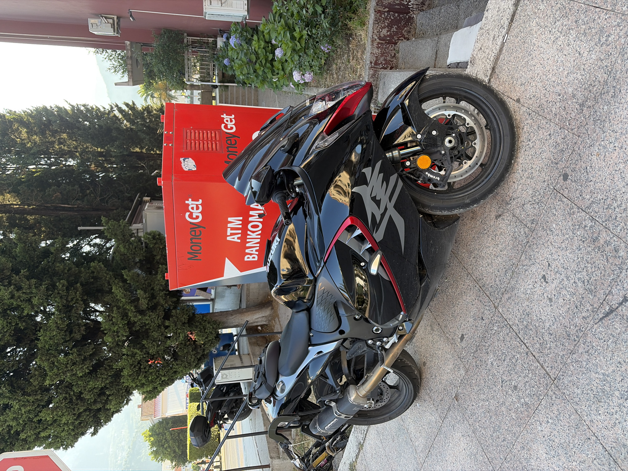 A black Suzuki Hayabusa motorcycle is parked on a tiled sidewalk in front of a red "Money Get" ATM kiosk. The ATM has "ATM BANKOMAT" written on it in white lettering. The motorcycle is positioned diagonally with its front wheel turned to the left. A black top box is mounted on the rear seat of the motorcycle. To the left of the ATM, another motorcycle is partially visible. A large evergreen tree is behind the ATM, and a building with a maroon facade is to the right. Lush green bushes with purple flowers are growing next to the building. The scene appears to be an outdoor urban setting, likely in Budva, Montenegro, given the presence of the ATM and the architectural style. The lighting suggests it is daytime.
