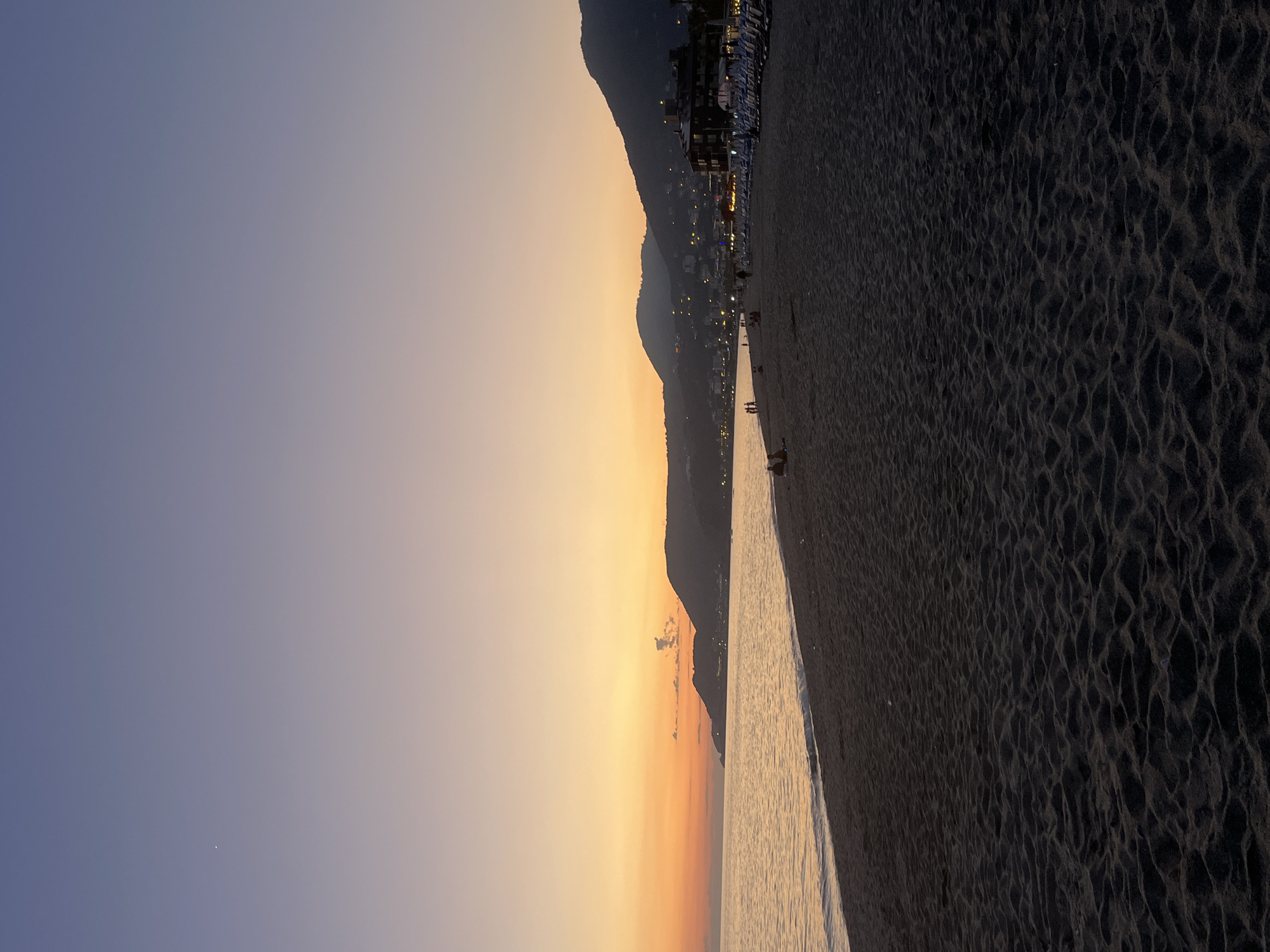 The image captures a coastline scene at dusk in Alanya, Turkey. A dark, sandy beach occupies the right half of the frame, its surface textured with wave patterns. Adjacent to the beach is a narrow strip of water, reflecting the ambient light. Beyond the water, the coastline is defined by a series of hills that rise upwards, dotted with buildings that glow with warm light, suggesting a developed area. The sky transitions from a deep blue on the left to a vibrant orange and yellow above the hills, indicating either sunrise or sunset. The light is dim, indicating time is around dusk or dawn. Some small figures are visible walking along the beach.