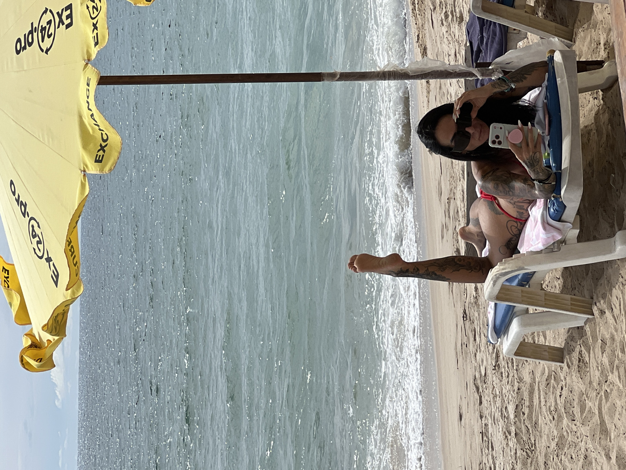 A woman with tattoos on her arms and legs lies on a beach chair on a sandy beach, facing the ocean. She is wearing a red bikini and sunglasses, and is holding a smartphone. A large yellow umbrella with the text "EX24.PRO EXCHANGE" is set up behind her, providing shade. The ocean is visible in the background, with waves breaking on the shore. The scene is set during the day, with a partly cloudy sky.