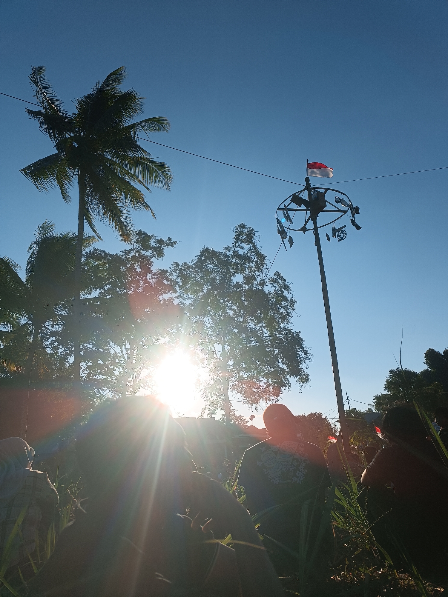 The image depicts a scene in Balearjo, Indonesia, under a clear blue sky. In the foreground, silhouetted figures are gathered amidst tall grass and foliage, partially obscured by the bright sun. The sun's rays create a lens flare effect, casting a glow over the scene. Behind the people, trees, including a prominent coconut palm on the left, are visible, with their leaves silhouetted against the sky.
A tall pole stands in the mid-ground, topped with a circular structure adorned with various small objects. At the very top of the pole, the Indonesian flag, red and white, is flying. Another Indonesian flag is visible in the far background, to the right of the pole. The overall atmosphere suggests an outdoor gathering or event, possibly a festival or commemoration, given the presence of the flags and the decorative pole. The low angle of the sun indicates it is either early morning or late afternoon.