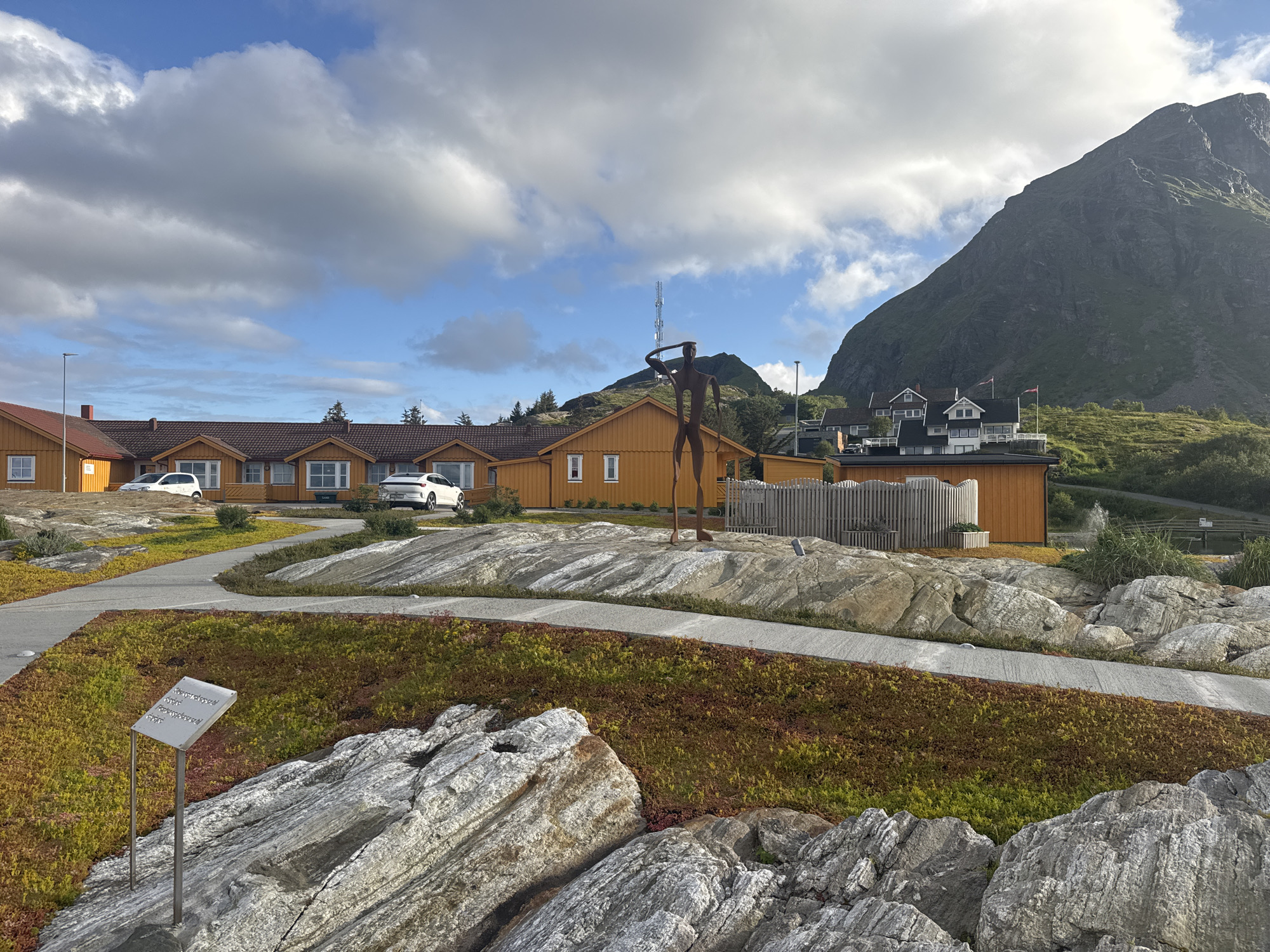 The photograph captures a scenic view in the city of Lovund, Norway, featuring a foreground of textured, light-colored rocks with patches of vibrant green and rust-colored ground cover. A metal signpost with informational text in an unfamiliar script stands prominently in the foreground. A concrete pathway winds through the rocky landscape, leading towards a cluster of modern, orange-colored buildings with dark roofs. In the middle ground, a stylized, tall, dark metal sculpture of a person stands, appearing to look out towards the horizon. Behind the sculpture, more orange buildings are visible, one with a wooden fence. A white vehicle is parked near one of the buildings, and another white car is visible further down the road. The background is dominated by a steep, grassy mountain slope with scattered houses and trees, under a sky filled with dynamic clouds and a clear blue sky. The overall impression is one of a well-maintained, modern development integrated into a rugged, natural environment.