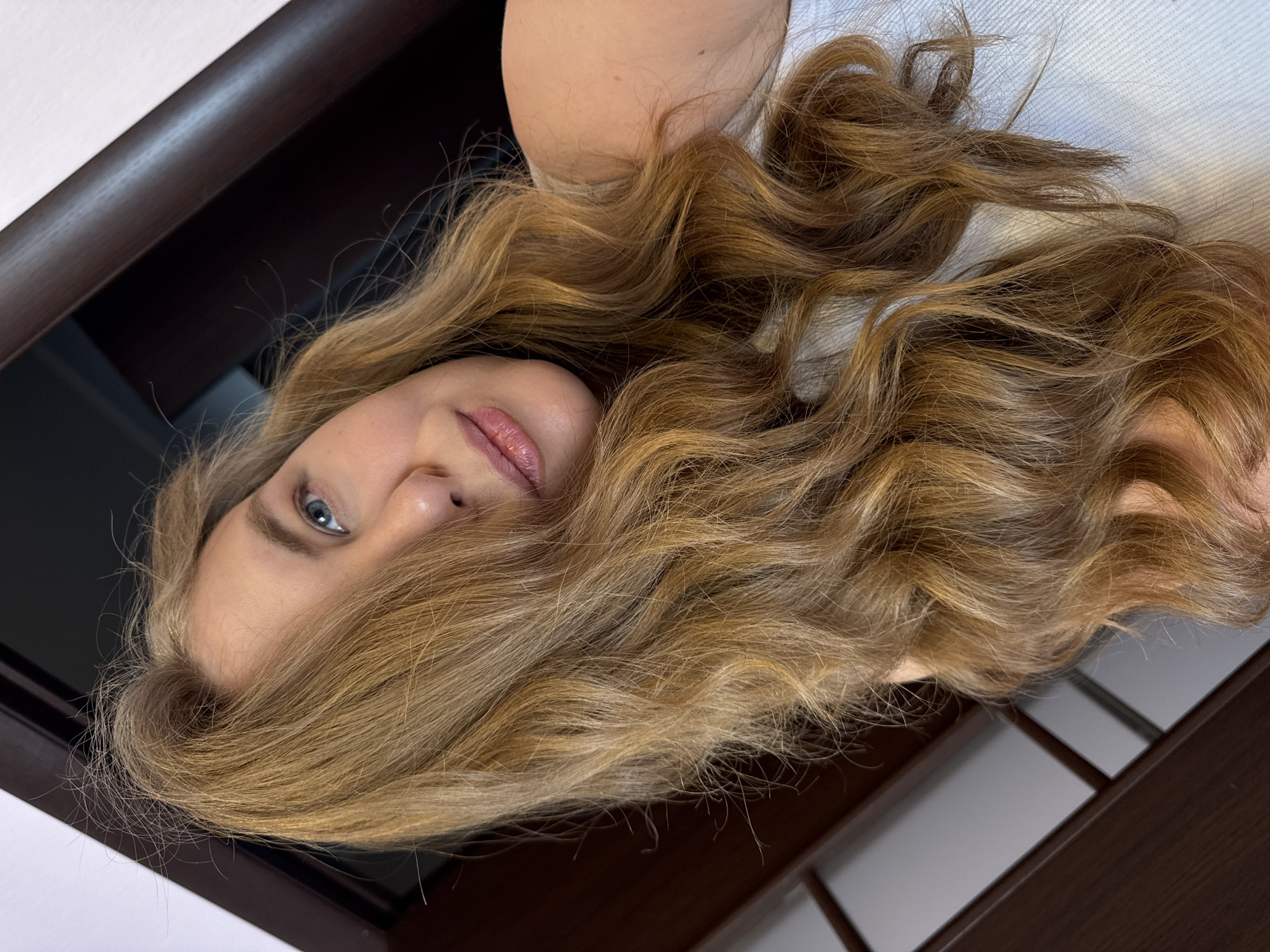 A close-up, low-angle selfie shows a blonde woman with long, wavy hair partially obscuring her face. She is looking upwards and to the left, with her blue eyes visible. Her lips are slightly parted and appear to be tinted pink. She is wearing a white top. The background is blurred, with dark brown wooden furniture and a white wall visible. The scene appears to be indoors.