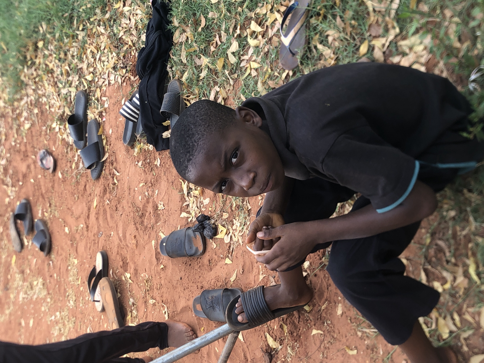 A young boy, appearing to be of African descent, crouches in an outdoor setting in Nsugbe, Nigeria. He wears a dark short-sleeved shirt with light blue trim on the sleeves and dark trousers, looking directly at the camera with a neutral, possibly solemn or curious expression. He holds a small, light-colored object in his hands, appearing to examine it. One of his bare feet rests inside a black slide sandal. The lower legs and bare feet of another person are partially visible on the far left, next to a metal bar or pipe.
The ground consists of reddish-brown earth and patches of green grass interspersed with numerous dry, yellowish-brown leaves. Scattered across this area are several pairs of footwear: multiple black slide sandals (some bearing a design similar to the "LV" logo, others with white stripes), a pair of black flip-flops with a tan sole, and a single brown flip-flop. A dark piece of clothing, possibly a shirt or jacket, is crumpled on the grass near some of the sandals. The scene depicts a casual outdoor moment in what appears to be a dry environment during daylight hours.