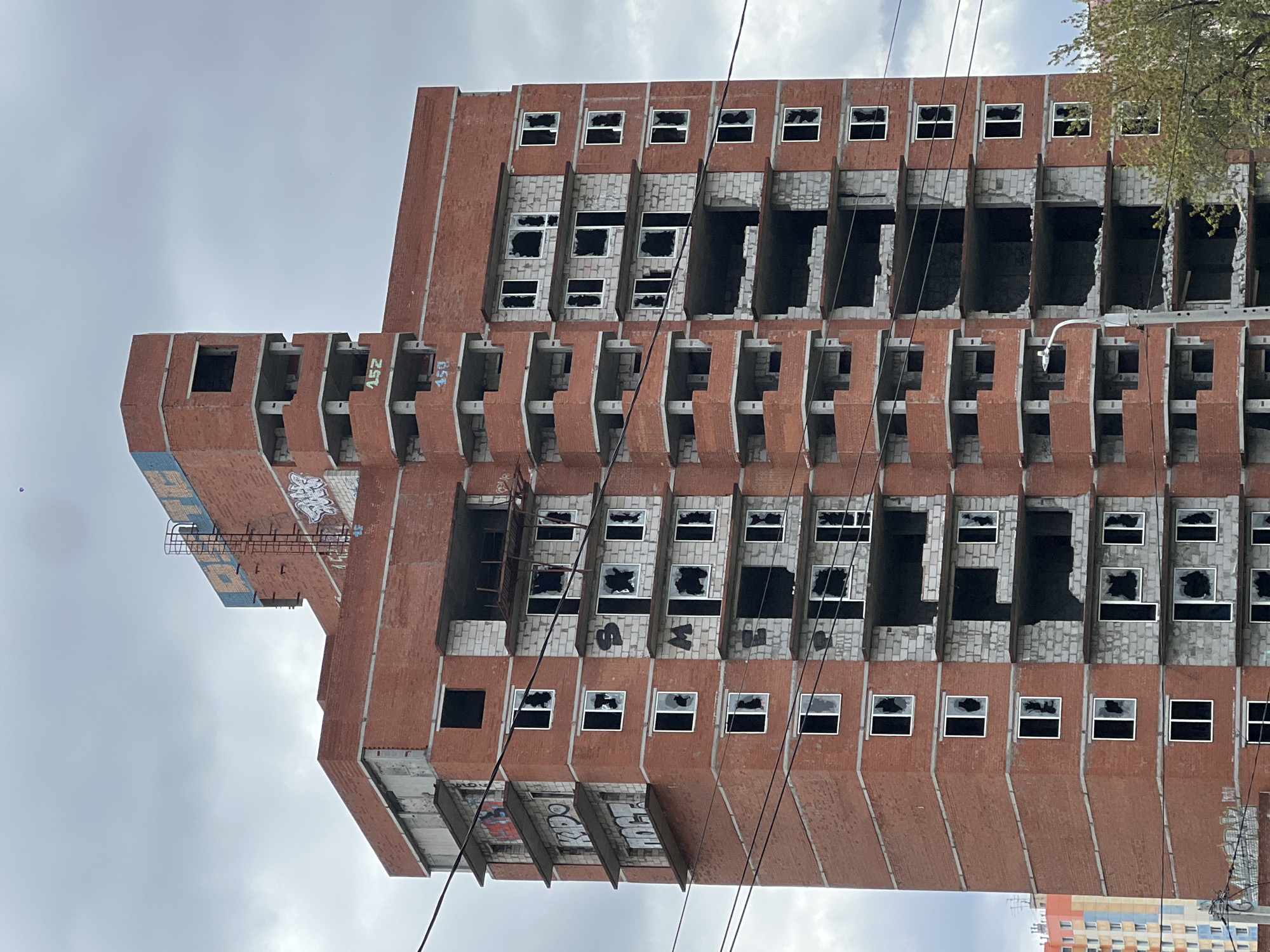 A tall, abandoned, brick building stands against a cloudy sky. The building is multi-storied with a modular design, each section featuring protruding balcony-like structures. Many of the windows are broken or missing. Graffiti is visible on the lower parts of the building. Power lines cut across the frame. The building is located in the city of Perm, Russia. Another building is visible in the bottom right corner of the frame. A small patch of tree leaves is visible in the top right corner. A ladder is visible on the side of the building.