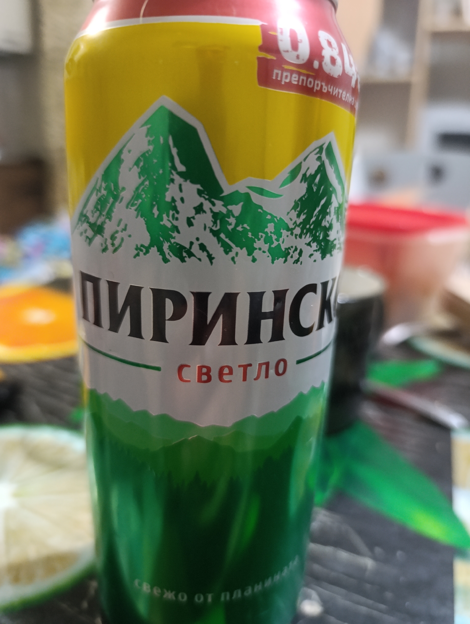 The image displays a close-up of a beverage can. The can has a yellow top portion with red accents, and features a graphic of green mountains with white snow caps. The main text on the can is in black capital letters, reading "ПИРИНСКО" (Pirinsko). Below this, in red lettering, it says "светло" (svetlo), meaning "light" in Bulgarian. Towards the top of the can, there is text that appears to be "0.8%" followed by "препоръчителна" (preporachitelna), which likely indicates recommended alcohol content. The bottom part of the can is predominantly green, with a textured pattern that suggests more mountains or natural scenery, and further text that reads "свежо от планината" (svezho ot planinata), meaning "fresh from the mountain." In the background, out of focus, there are some kitchen-like elements, including a red container and what appears to be a sliced lime or lemon on the left. The overall scene is likely a domestic setting, possibly a kitchen counter. There are no people or specific landmarks visible that would indicate a precise location within Kyustendil, Bulgaria, other than the brand name itself, which is associated with the Pirin Mountains in Bulgaria.