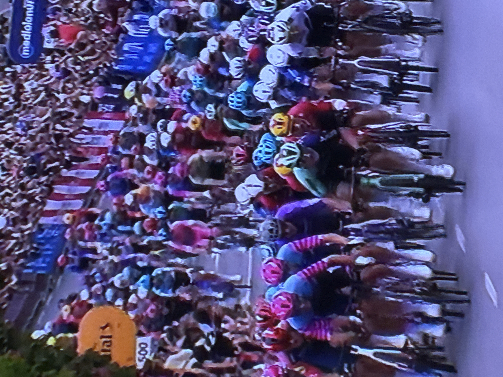 The image shows a large group of cyclists racing on a road, surrounded by a crowd of spectators. The cyclists are wearing helmets and brightly colored jerseys. The crowd is standing behind barriers lining the road. A yellow sign with "tal s" and "500" is visible. A Mediolanum sign is also visible. The location is most likely not in Sofia, Bulgaria, as nothing in the image suggests it.