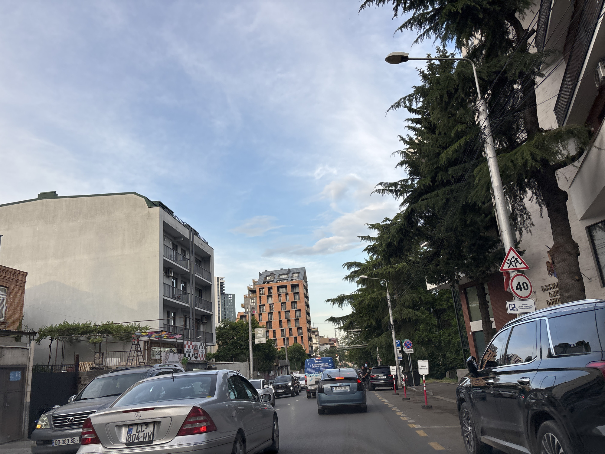 This is a street-level view captured from a vehicle moving through Tbilisi, Georgia. The scene depicts a moderately busy street with several cars, including a silver Mercedes-Benz with license plate 804-WW and a silver Lexus RX with license plate DD-080-BB. The buildings lining the street vary in style and age. On the left, there's a light-colored, multi-story building with balconies, adjacent to a smaller brick structure. Further down the street, a striking, orange brick building with numerous windows stands out. Tall, dense evergreen trees line the right side of the road, interspersed with a light pole and various street signs, including a "40" speed limit sign and a pedestrian crossing warning. The sky above is partly cloudy with a mix of blue sky and white clouds. The presence of modern apartment buildings and traffic suggests an urban environment within Tbilisi.