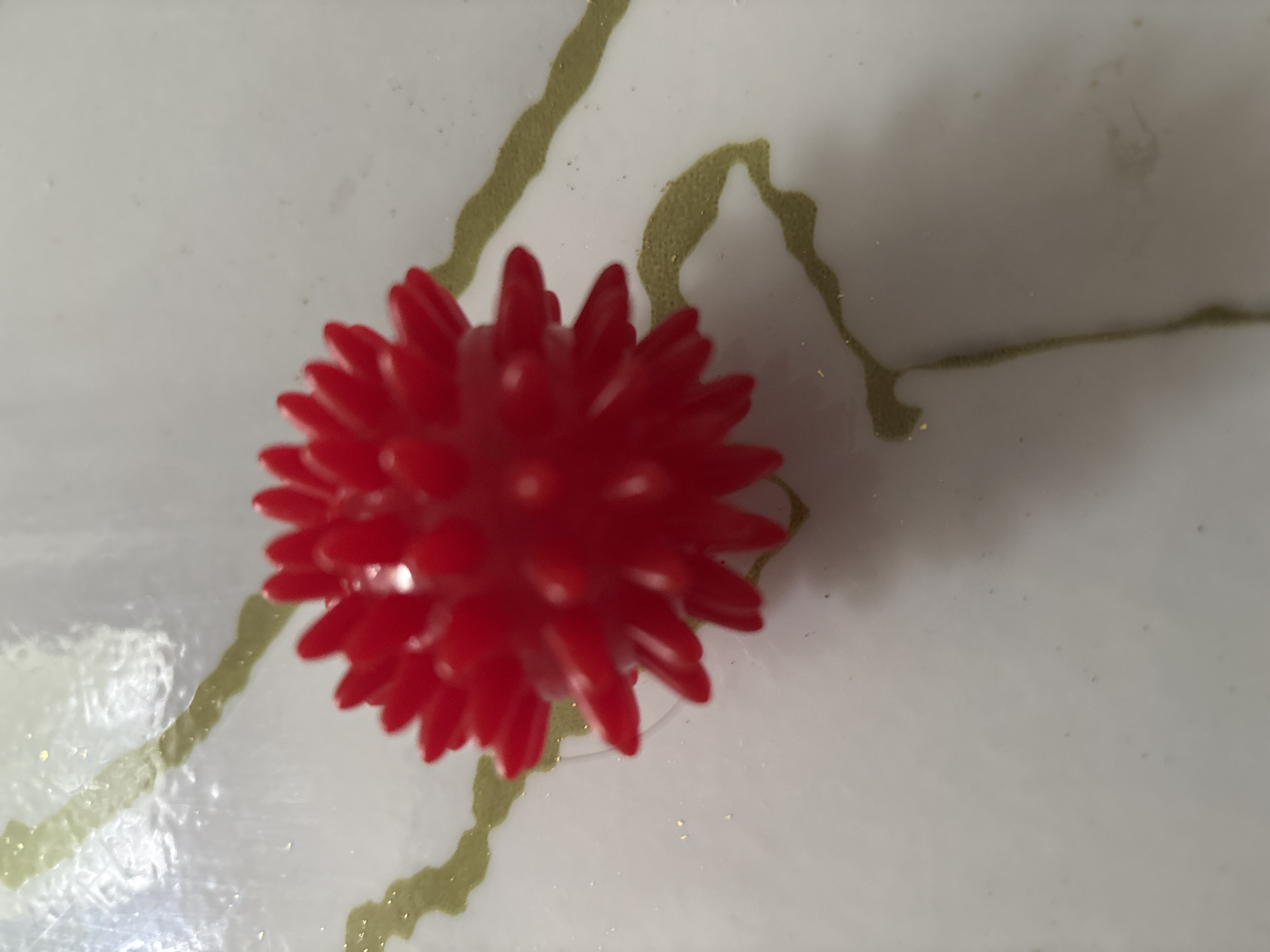 A close-up, overhead shot focuses on a spiky red ball, resembling a stress ball or a toy, positioned on a white surface. The surface appears to be marbled with thin, irregular streaks of a golden-greenish hue, possibly paint or a decorative element. The lighting is soft, with subtle shadows cast by the object, suggesting an indoor setting. There is no visible text in the image. The overall composition is simple, emphasizing the texture and color of the red object against the patterned background.