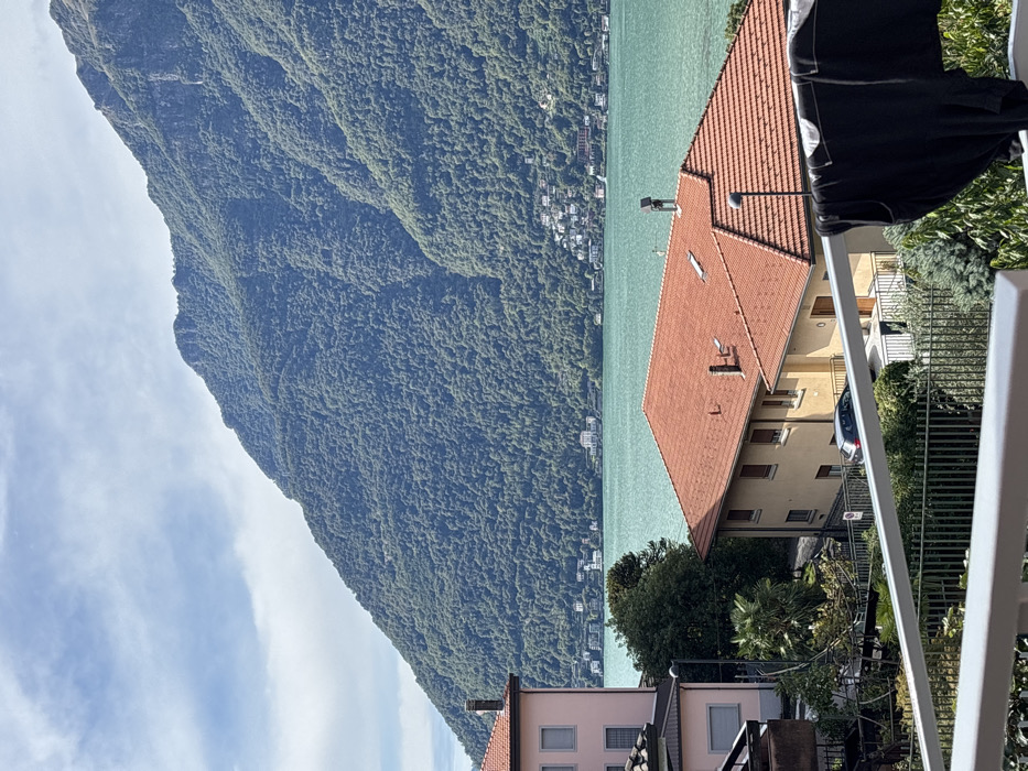 The image displays a panoramic view of Lake Como from Nesso, Italy, under a partly cloudy sky. A large, verdant mountain, densely covered in deciduous trees, dominates the background, rising steeply from the turquoise-colored lake water. Along the distant shoreline on the opposite side of the lake, numerous white and light-colored buildings are visible at the mountain's base.
In the midground, a multi-story building with a terracotta-tiled roof and pale yellow walls occupies the center-right, featuring multiple windows with closed brown shutters. A small, dark rectangular object, possibly a solar panel or vent, is positioned on its roof. To the left, a partially obscured pink-hued building with a similar roof is visible. Various green trees and foliage, including a prominent palm tree, are interspersed between and around the buildings. A dark-colored hatchback vehicle is parked on a paved surface or road behind a dark green metal fence.
The scene is framed from a balcony, indicated by a white railing extending across the bottom of the image. Black clothing, specifically a dark t-shirt or similar garment, hangs drying on a rack in the lower right foreground. No individuals are present in the image.