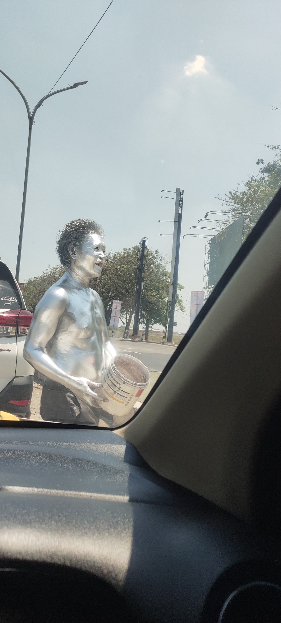 A person covered in silver paint stands beside a road in Bolo Krajan, Indonesia, holding a partially filled bucket of paint. The person's hair is also silver, and they appear to be an adult. They are wearing dark pants. The scene takes place outdoors during the day, with a bright sky and scattered clouds visible. Trees and utility poles line the road, and a white car is parked nearby. The image is taken from inside another vehicle, with the dashboard and a portion of the windshield visible in the foreground.