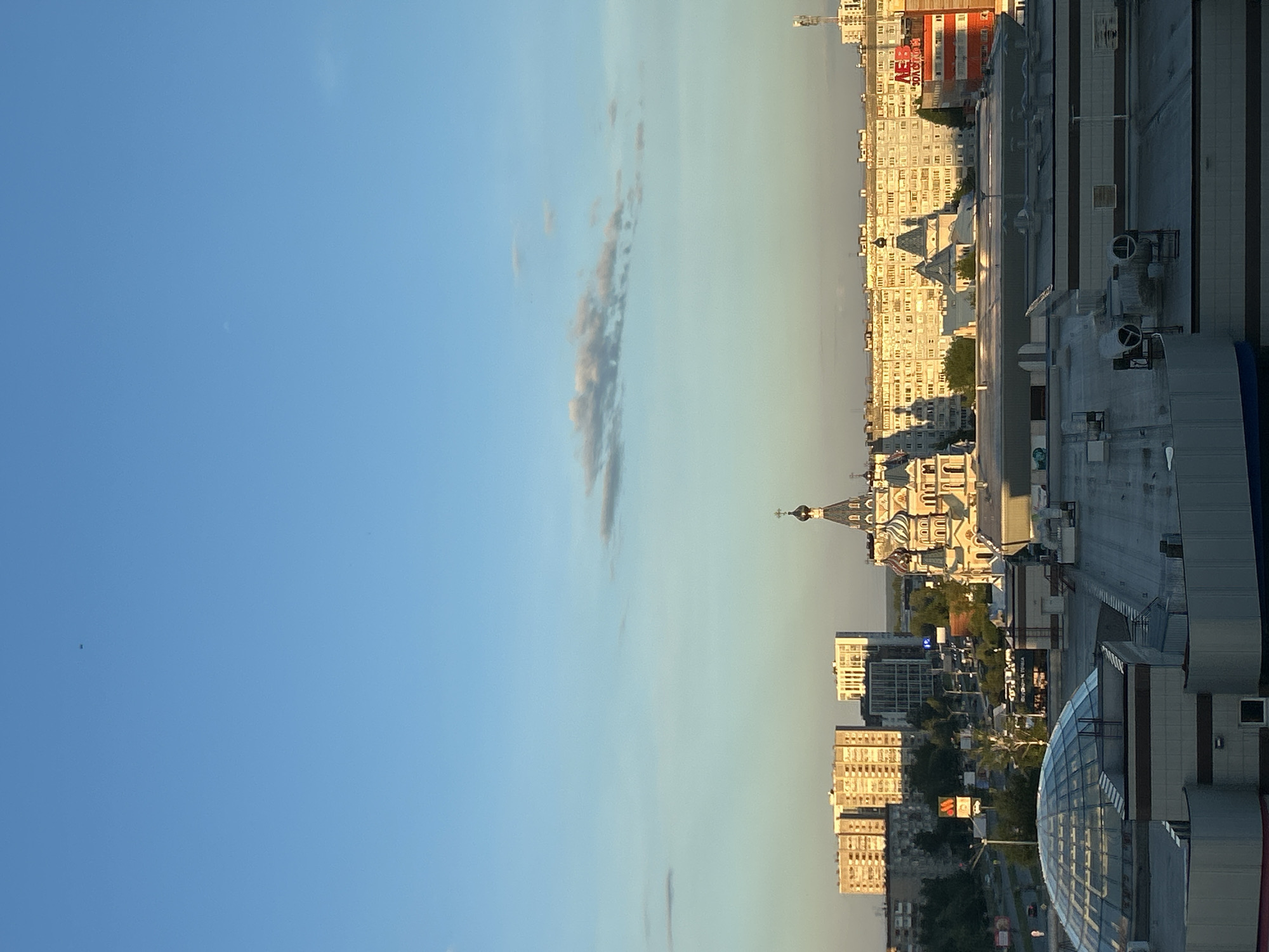 The image displays an urban panoramic view of Izhevsk, Russia, captured during the golden hour, likely late afternoon or early evening, as indicated by the warm, directional light casting long shadows. The sky is predominantly clear blue, transitioning from a lighter hue near the horizon to a deeper blue higher up, with a distinct, elongated cloud formation stretching across the middle ground.
The scene is dominated by a dense collection of city buildings, varying in architectural style and height. A prominent feature is a large, ornate building resembling a church, characterized by multiple domes, some with onion shapes, and topped with crosses, consistent with Russian Orthodox architecture. Numerous multi-story residential and commercial buildings are visible, bathed in the warm sunlight, with some exhibiting signs of wear or older construction.
In the foreground and to the right, a modern building with a large, curved glass roof is visible, suggesting a shopping mall or a public complex. Amidst the urban sprawl, pockets of greenery from trees can be seen.
No people are explicitly visible in the wide shot or the provided crops.
Notable details include:
* **Time of day:** Golden hour, evident from the warm light and long shadows.
* **Weather:** Clear blue sky with scattered, wispy clouds.
* **Location cues:** The onion-domed church is a strong indicator of Russian architecture. The provided context specifies Izhevsk, Russia.
* **Visible text:** On a red and white building, the Cyrillic text "АЕВ" and "ЭЛБРУС" is discernible. On a yellow sign, likely "ВКУСНО и ТОЧКА" (Vkusno i tochka), a Russian fast-food chain, can be seen.