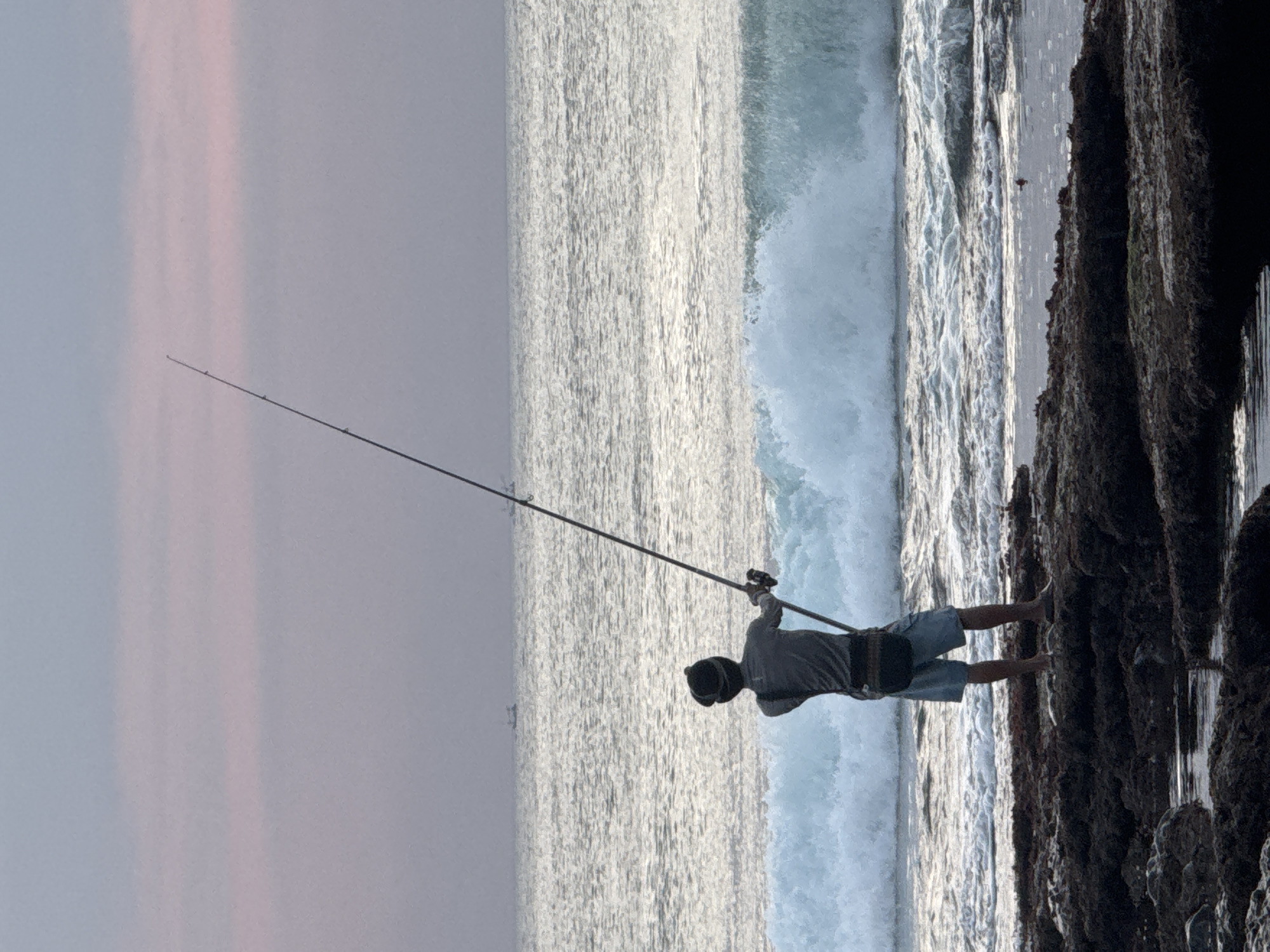 A lone fisherman stands on a rocky shore in Kuta Selatan, Indonesia, casting a fishing rod into the ocean as waves crash behind him. The sky above is a soft gradient of pink and purple, suggesting either sunrise or sunset. The ocean is a shimmering expanse of grey and white, with a large wave breaking in the mid-ground. In the distance, two small boats can be seen on the horizon. The fisherman is wearing a grey shirt, blue shorts, and a dark cap. He is holding the fishing rod with both hands, his body angled towards the sea. The rocks in the foreground are dark and textured, with some pools of water reflecting the sky. The overall impression is one of solitude and peaceful contemplation amidst the raw beauty of the natural environment.