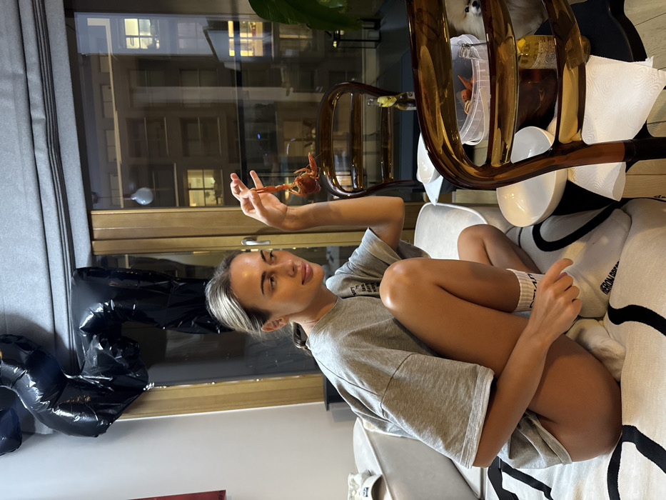 A woman in a gray t-shirt sits on a sofa, legs tucked, gazing at a crawfish she holds in her hand. The crawfish is positioned above her outstretched hand, close to a window showing an urban night scene. Behind her, black balloons decorate the room. Nearby, a brass-colored object resembling musical instrument components sits on a table, also featuring dining ware and napkins. The room appears to be an apartment with modern decor.