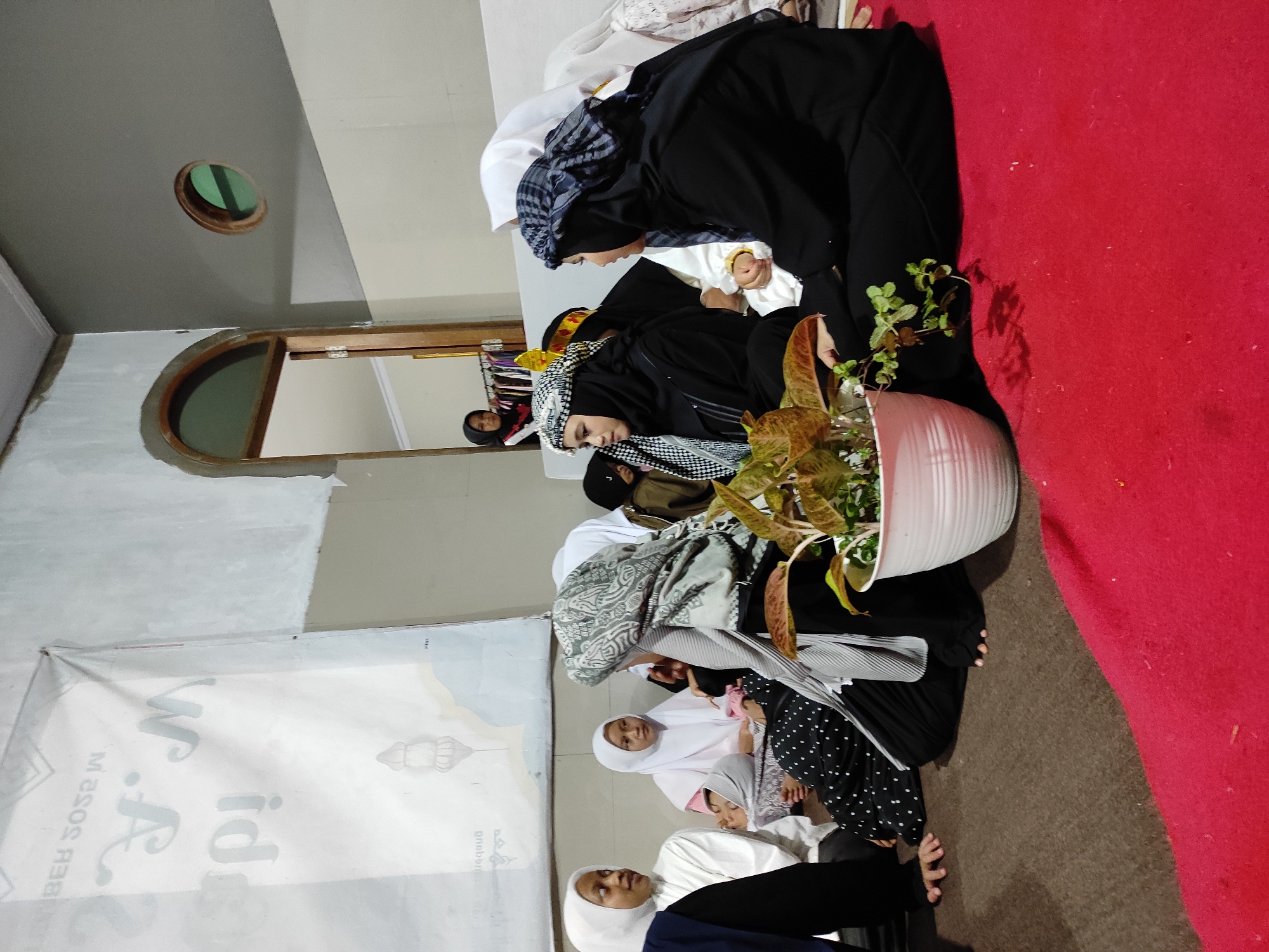 In Sumedang, Indonesia, a group of Muslim women and girls are gathered indoors, likely for a religious or cultural event. The scene is set on a floor covered with brown carpet and a vibrant red rug. A white pot with a leafy plant sits prominently in the foreground. Several women and girls are seated on the floor, many wearing hijabs and traditional Islamic clothing. One woman, on the right, is wearing a black abaya and a checkered headscarf. Another woman next to her, also in black, has a white hijab. In the background, through an arched doorway, another young woman in a hijab can be seen. A banner with Arabic text and an illustration of a lantern hangs on the wall to the left. The room has plain walls and a simple ceiling with a circular vent. The lighting appears to be artificial, creating a warm ambiance.