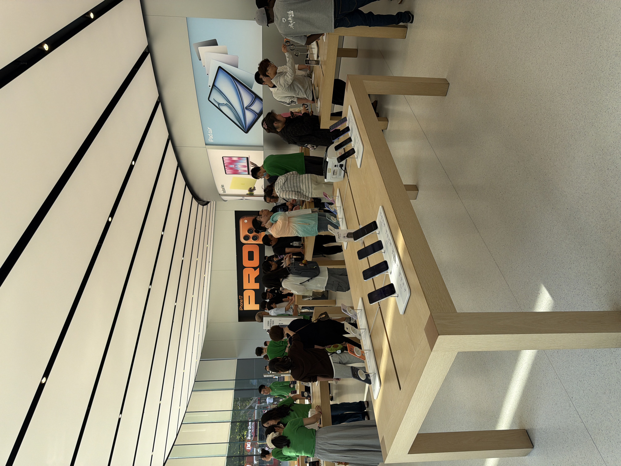 Inside an Apple store in Xuanwu District, China, a long wooden display table showcases iPhones. People are gathered around the table examining the devices. A large "PRO" display and iPad Air advertisements are visible in the background. The store has a high ceiling with black and white paneling. Large windows line the wall on the left.