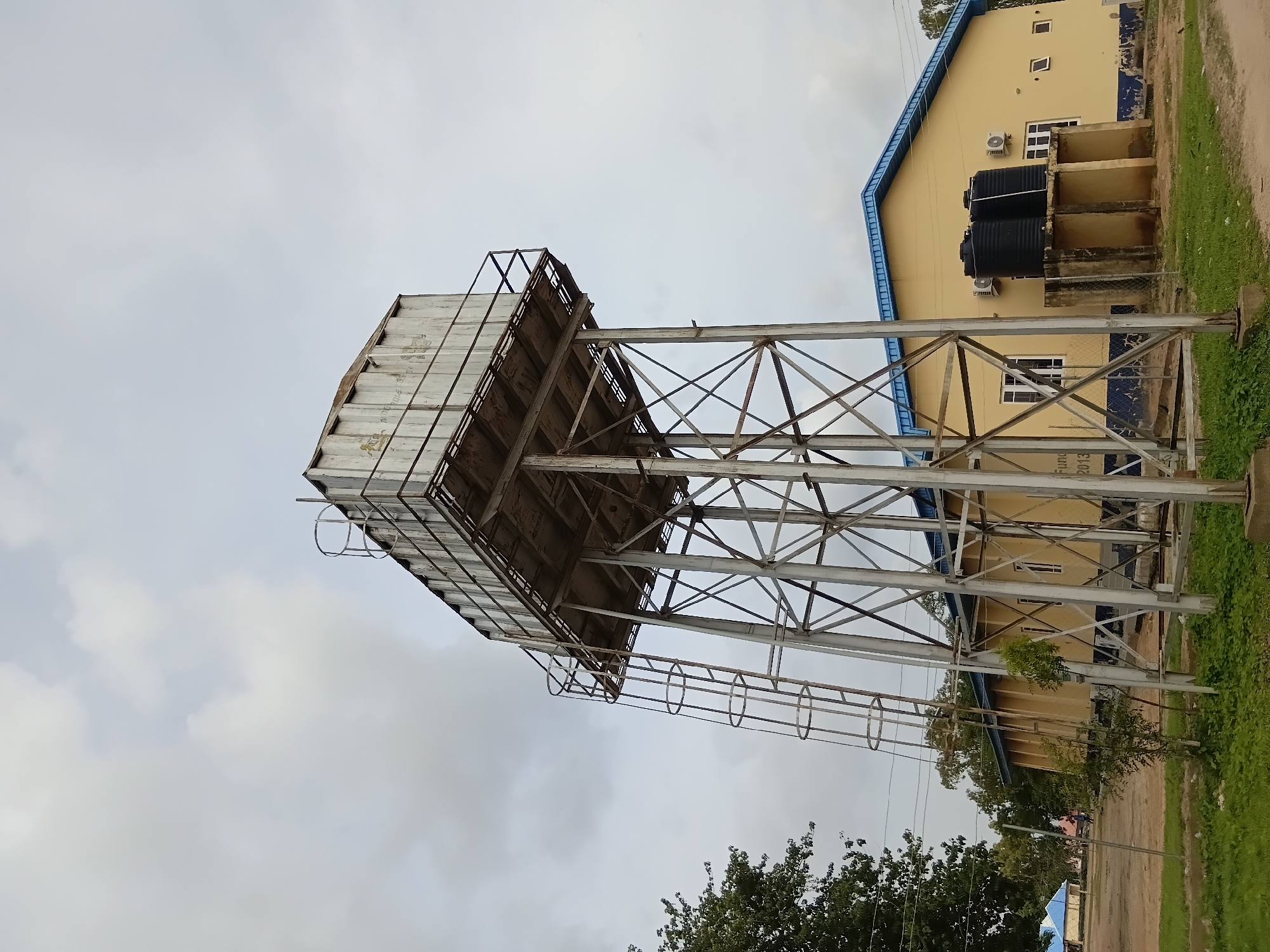 This image shows a water tower and a building in Jalingo, Nigeria. The water tower is made of metal and has a ladder on its side. The building is yellow with a blue roof and has two air conditioning units on its exterior. There are also two black water tanks on a concrete platform next to the building. The scene appears to be outdoors, with trees and grass visible in the foreground and background. The sky is cloudy. The overall impression is one of a functional, possibly public, facility.