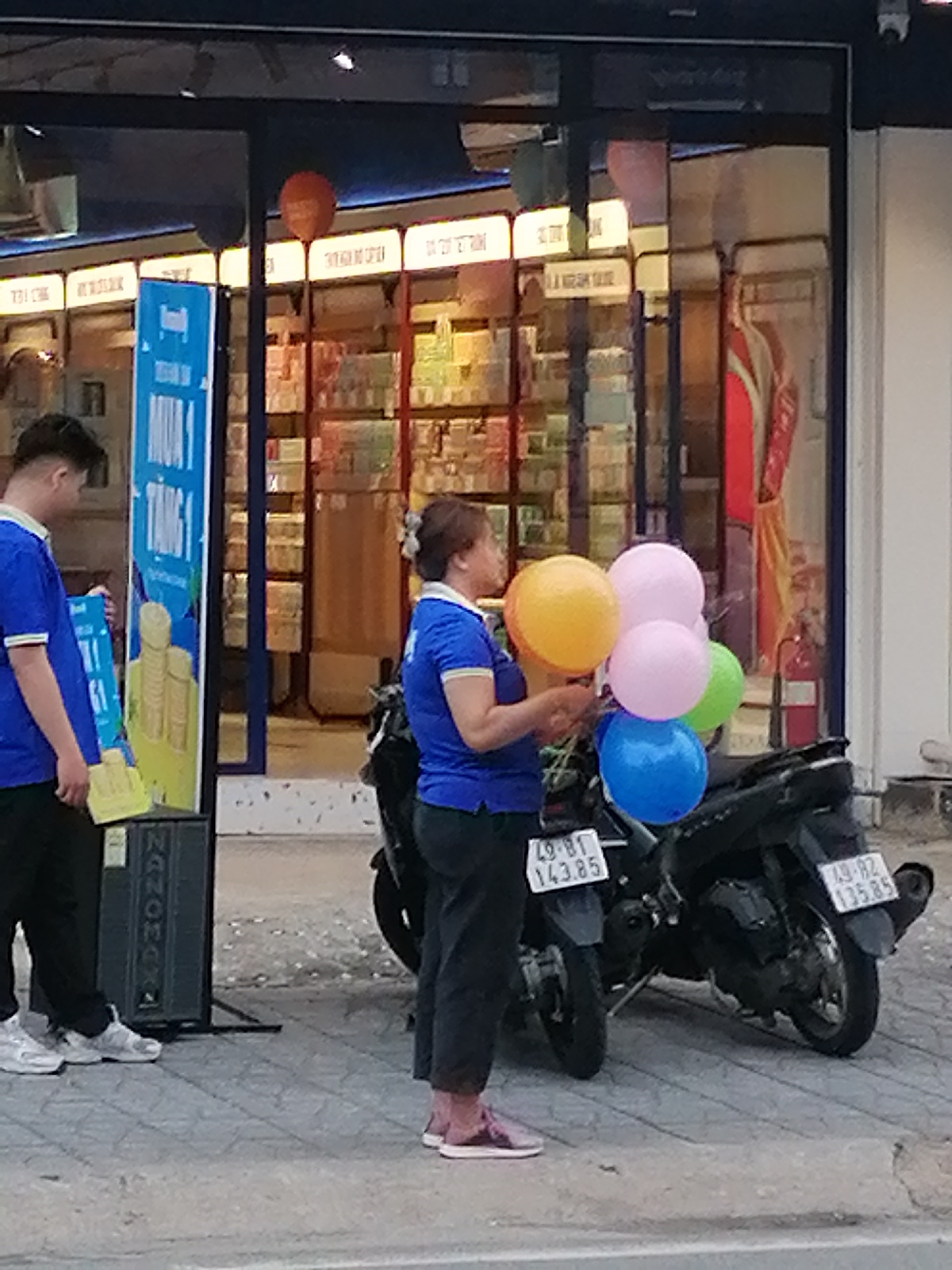 The photo was taken in what is possibly Da Lat, Vietnam. An Asian woman in a blue polo shirt and dark pants holds five balloons of different colors: yellow, pink, light green, and blue. The woman is standing next to a black motorbike with license plates reading "49-B1 143.85" and "49-B2 135.85". A tall blue sign with Vietnamese characters is set up to the left of the woman, and a man in a matching blue polo shirt is standing next to the sign. A gray speaker is next to the man and sign. The background features a store front with glass windows revealing shelves stocked with merchandise. An orange balloon is visible near the top of the frame.