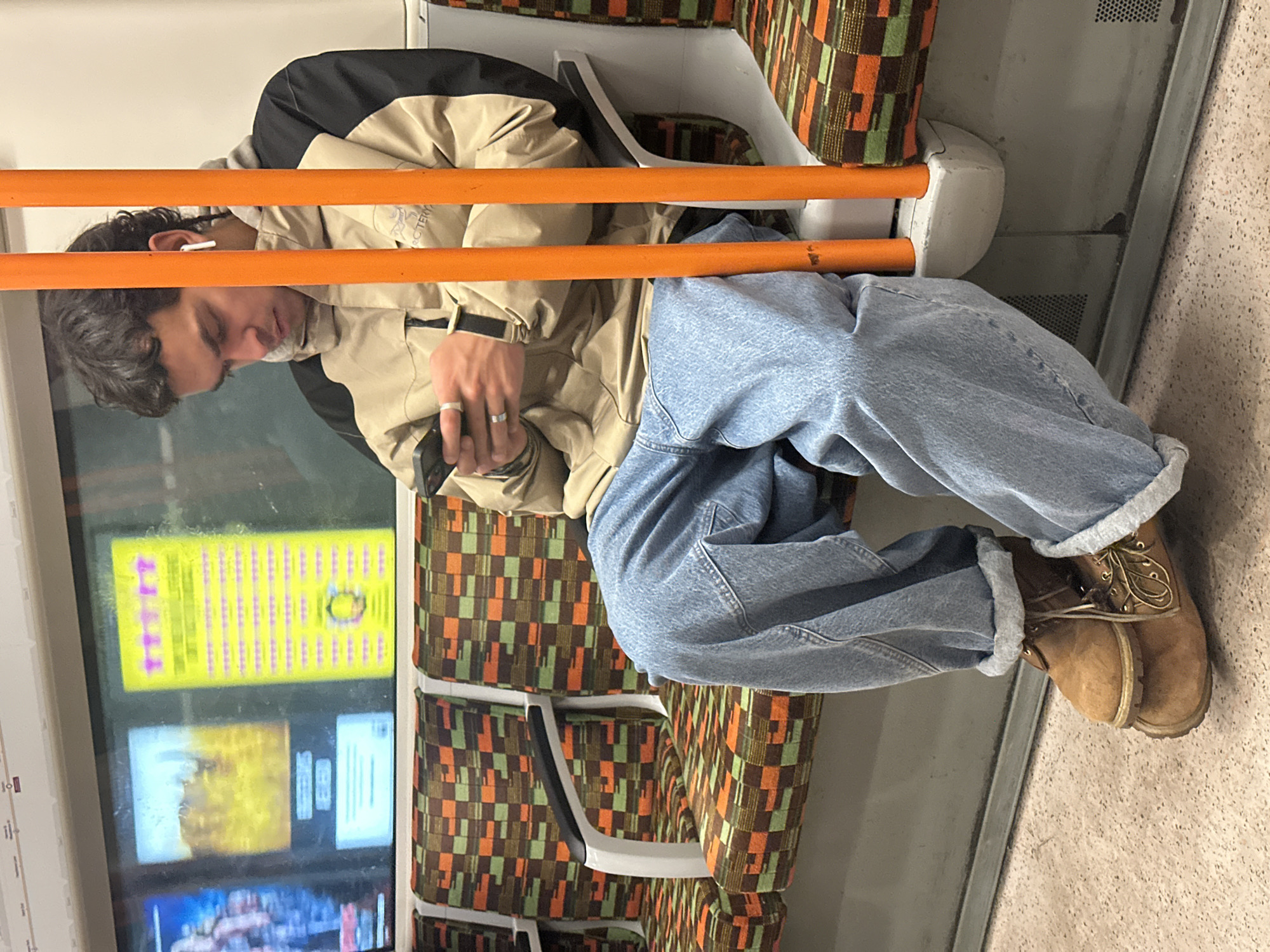 A young man is seated on the London Underground, likely on the Circle, District, Hammersmith & City, or Metropolitan line, as indicated by the orange grab rails. He is wearing a beige jacket, light-wash denim jeans cuffed above his ankles, and tan work boots. He has an earbud in his left ear. His right hand is holding a smartphone. The background features the train's windows displaying advertisements, patterned seating typical of the Underground, and interior train details.