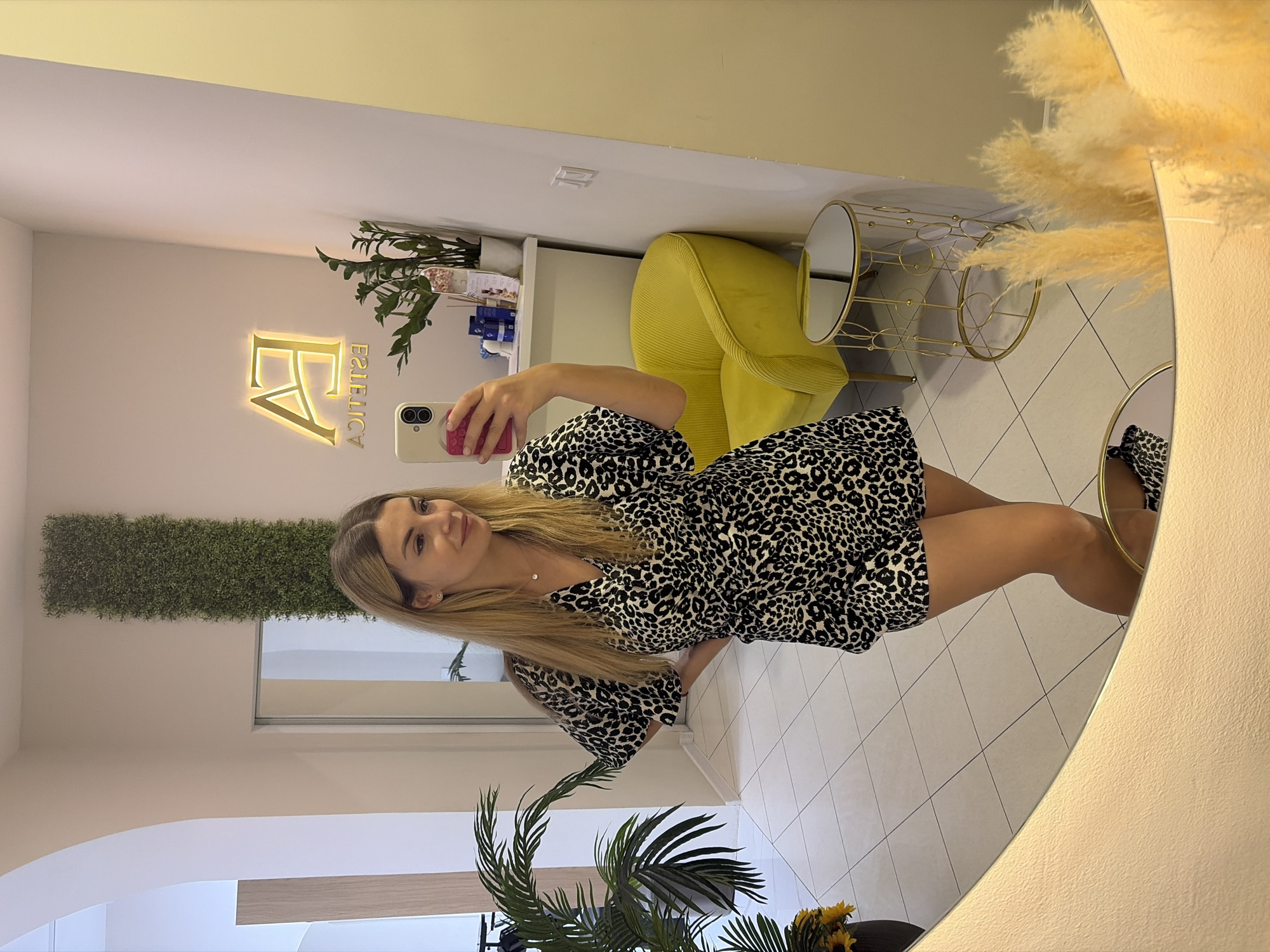 The image shows a woman taking a selfie in a mirror. She is standing in a well-lit room with white tiled floors. The woman has blonde hair and is wearing a black and white leopard print dress. She is holding a smartphone in her right hand, and her left hand is on her hip. In the background, there is a bright yellow armchair, a small round golden table, and a neon sign on the wall that reads "EA ESTETICA". There is also a tall vertical arrangement of artificial greenery on the wall. The room appears to be a salon or spa. The woman's reflection is visible in the mirror, along with parts of the room's interior. Based on the visible elements and typical salon decor, this location is likely in Budva, Montenegro.