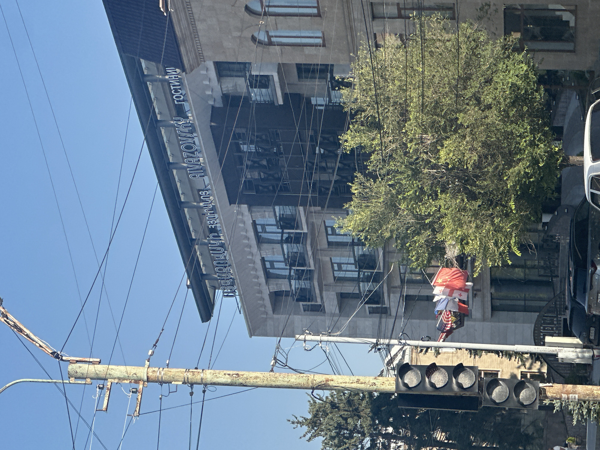 A street view in Gyumri, Armenia, shows a building with "AIVAZOVSKY" and "ГОСТИНИ" (meaning "hotel") written in illuminated letters on its facade. A traffic light pole with multiple signal lights is on the left, with wires crisscrossing the scene. Several flags, including the US, Canadian, and Georgian flags, are hanging from wires on the traffic light pole. A lush green tree is situated in front of the building. The sky is clear and blue.