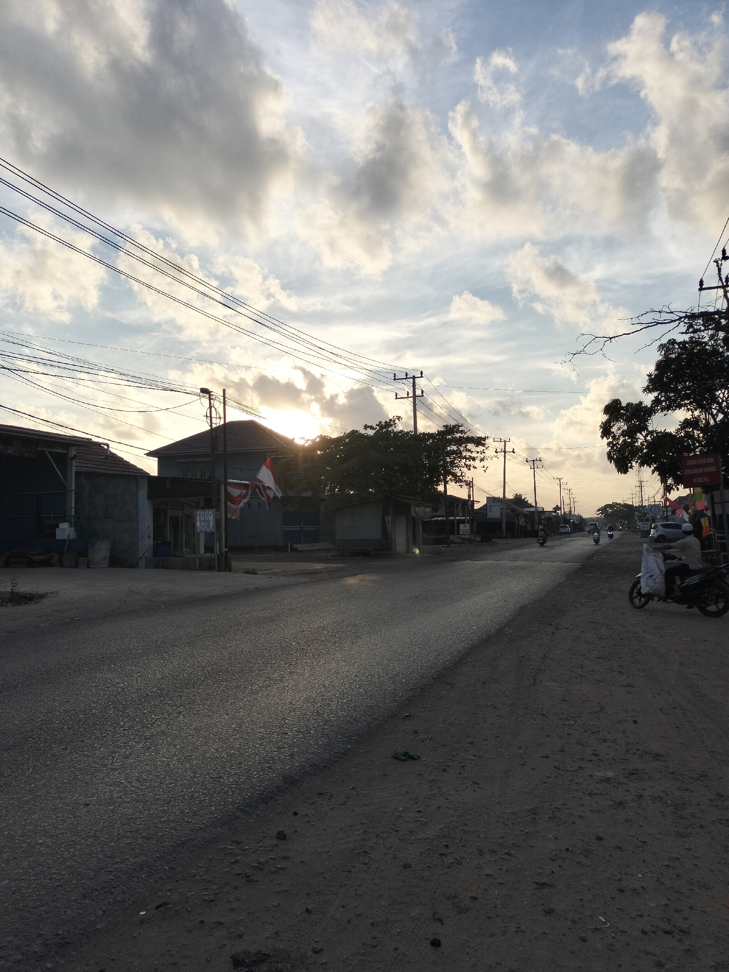 The image shows a street scene in Karang Indah, Indonesia, during what appears to be late afternoon or early evening. The sun is low in the sky, casting long shadows and creating a warm, diffused light. The sky is filled with scattered clouds, with the sun breaking through in the center-left of the frame, partially obscured by them.
The street itself is paved and appears to be a main thoroughfare, with a paved section on the left and a dirt shoulder on the right. Several utility poles with overhead wires line the street. Along the left side of the road, there are buildings that appear to be shops or small businesses, one of which has a sign with Indonesian text. There are two flags flying in front of one of the buildings: the Indonesian flag and another red and white flag.
On the right side of the road, more businesses are visible, including one with a sign that appears to be for a shop or market. Several motorcycles are present, with one rider in the foreground on the right, carrying a large white bag. Other motorcycles and a car are visible further down the street. Trees are present on both sides of the road, with some bare branches visible against the sky. The overall impression is one of a typical Indonesian street scene, with signs of daily life and commerce under a dramatic sky.