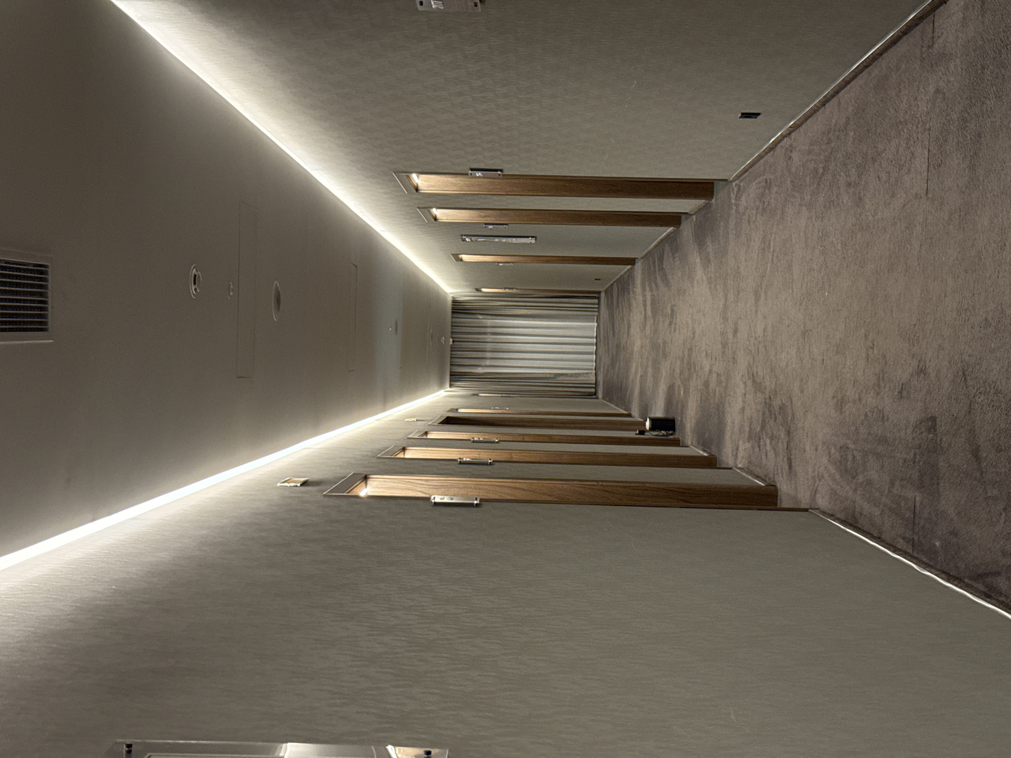 The image shows a long, dimly lit hotel hallway. The hallway features a textured wallpaper on the walls, a plush brown carpet on the floor, and a linear LED light fixture running along the top of the walls on both sides. Several closed wooden doors with metal door handles are visible along the right side of the hallway, each with a small rectangular sign above it. On the left side, the wall is mostly plain, with a few recessed lights and a vent visible on the ceiling. At the far end of the hallway, striped curtains are drawn. The scene is sterile and appears to be unoccupied. The location is identified as Kerch, Ukraine.